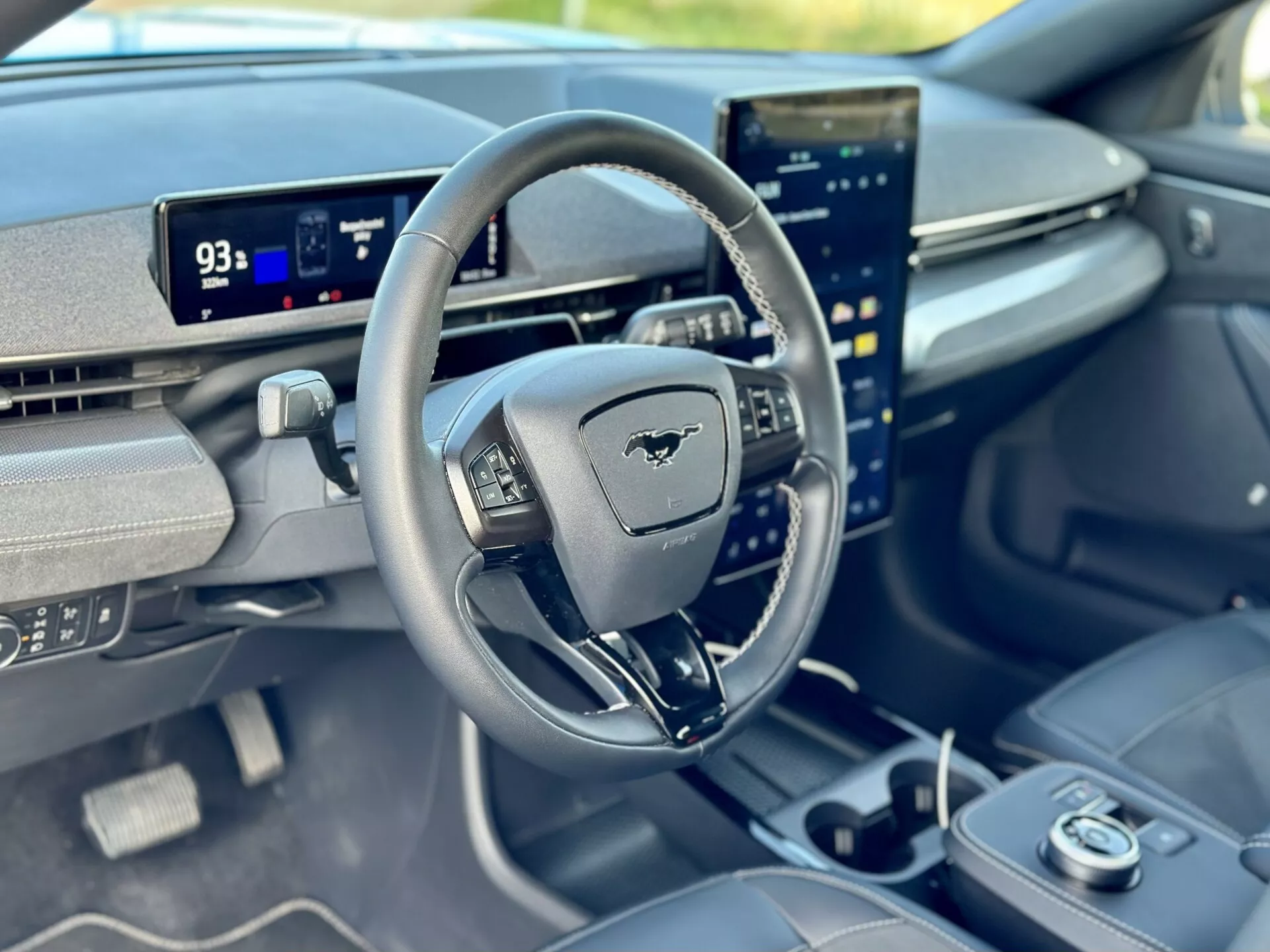Widok wnętrza Ford Mustang Mach-E 2020 skupiający się na kokpicie kierowcy z perspektywy pasażera przedniego, prezentujący kierownicę z emblematem Mustang, cyfrowy deska rozdzielcza w trybie parkowania pokazująca zasięg 93 mil, i pionowo zorientowany centralny ekran dotykowy zintegrowany z zaawansowanymi możliwościami systemu infotainment.
