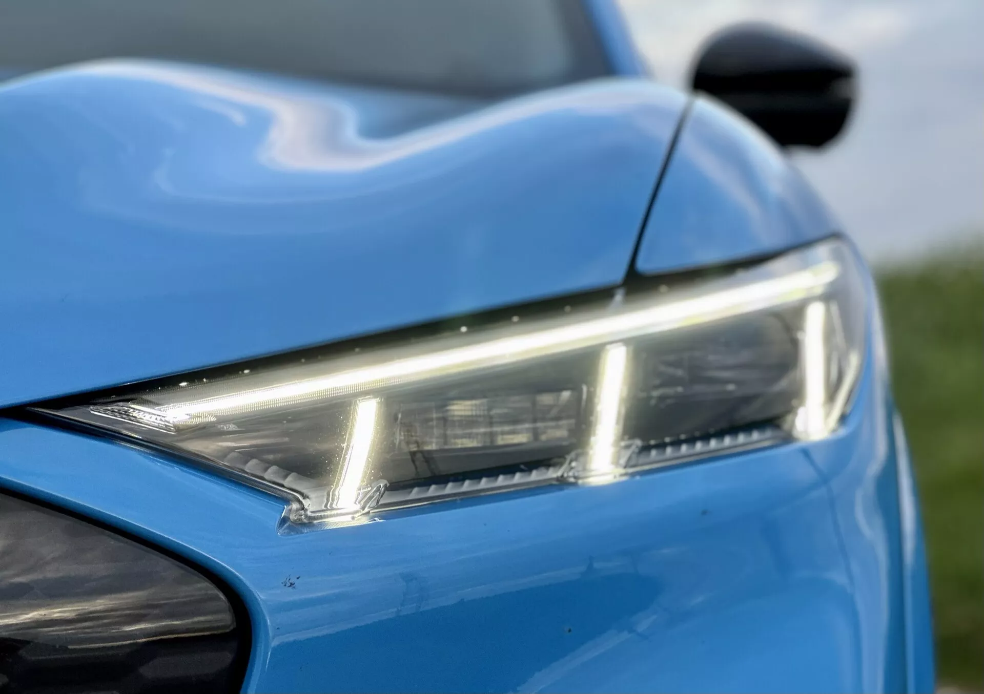 Zbliżenie na przednią lewą część Forda Mustang-Mach-E 2020 ze szczegółowym widokiem eleganckiego reflektora LED, dynamicznych linii nadwozia w żywym niebieskim kolorze, z widocznością części zderzaka i lusterka bocznego, podkreślające nowoczesny design i estetykę.