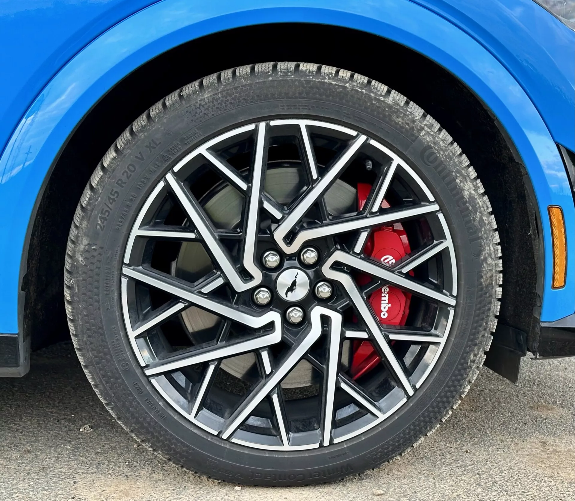 Zbliżenie na przednie prawe koło Forda Mustang Mach-E 2020 z wieloramiennej felgą ze stopu i wysokowydajnym układem hamulcowym z czerwonym zaciskiem, ukazujące szczegóły ogumienia całorocznego oraz fragment intensywnie niebieskiego błotnika, zdjęcie z niskiego kąta.