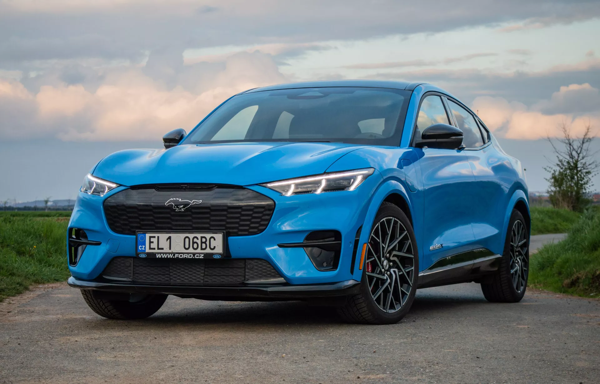 Niebieski Ford Mustang Mach-E z 2020 roku sfotografowany pod niskim i nieco ekscentrycznym kątem, prezentujący grill, reflektory, maskę i profil SUV-a, w tym przednie koło i błotnik, podkreślając jego wydajność i aerodynamiczny design o świcie lub zmierzchu.