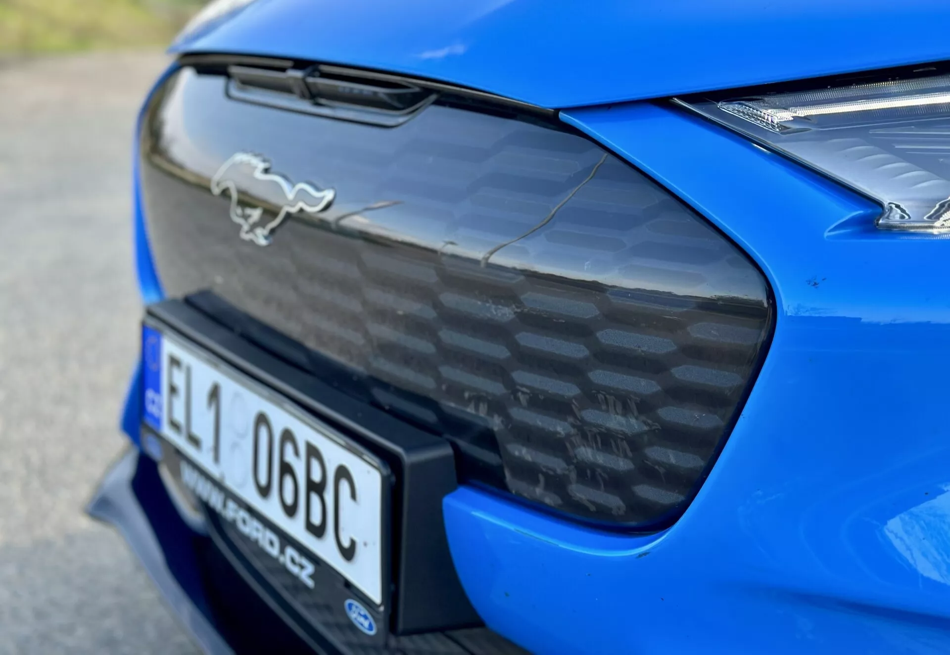 Zbliżenie na tylną część niebieskiego sportowego samochodu Ford Mustang Mach-E 2020 z chromowanym emblematem konia, tylnym spojlerem z włókna węglowego, zintegrowanym miejscem na tablicę rejestracyjną i częściowym widokiem na tylne światła LED, podkreślające aerodynamiczne i wydajnościowe cechy samochodu.