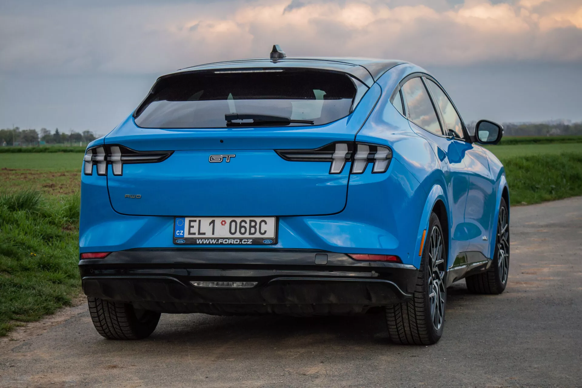 Ford Mustang-Mach-E 2020 sfotografowany z tylnego trójkątnego kąta, ukazujący jego charakterystyczny design, w tym poziome tylne światła LED, emblemat producenta, zintegrowany spoiler tylny, zderzak tylny wyposażony w sensory bez widocznego wydechu sugerujący status pojazdu elektrycznego, europejską tablicę rejestracyjną, wieloramienne felgi ze stopu, żywą niebieską farbę i aerodynamicznie ukształtowaną karoserię, na tle wiejskim.