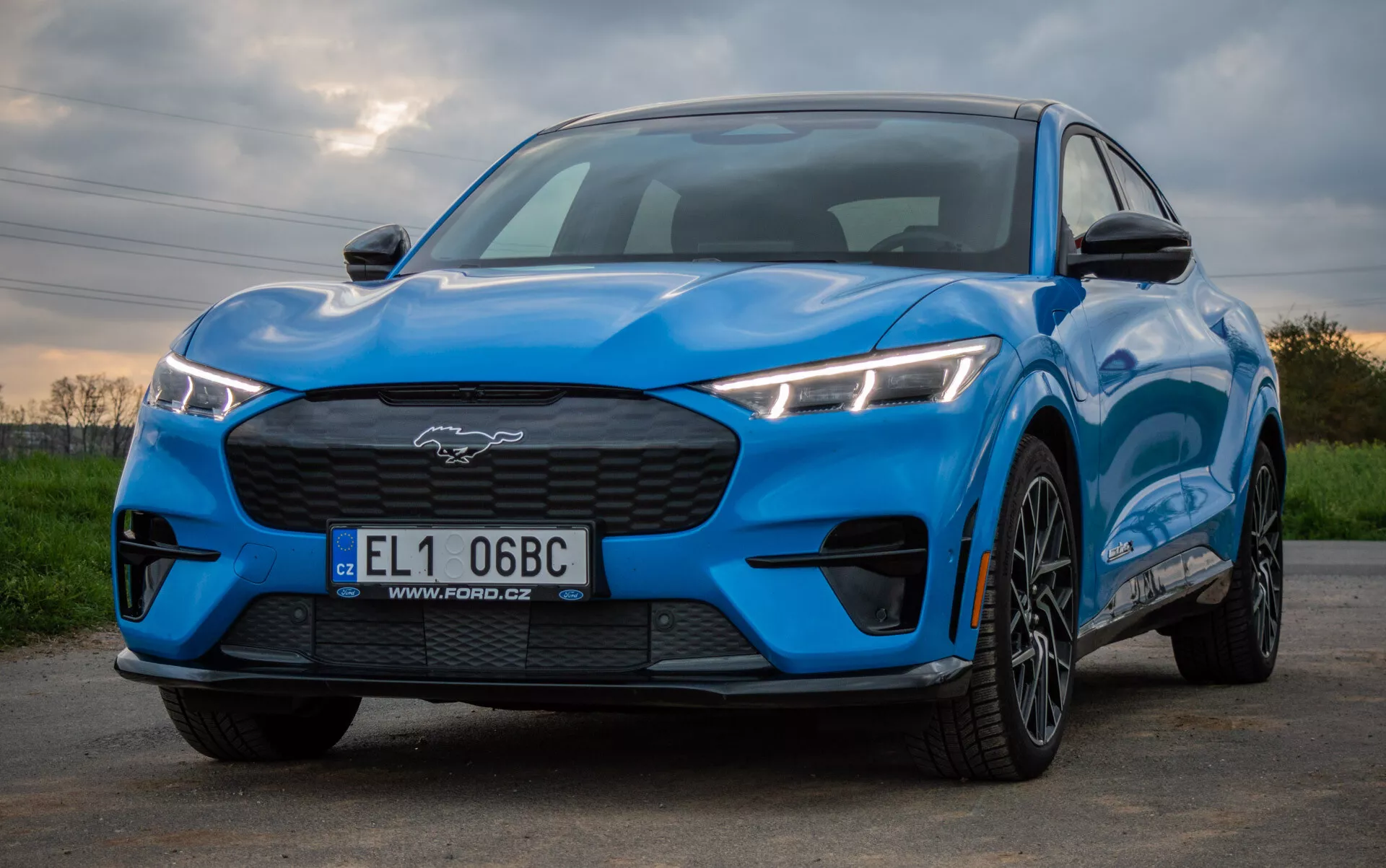 Ford Mustang Mach-E 2020 w jasnym niebieskim kolorze prezentujący sportowy i aerodynamiczny design z niskiej perspektywy przednio-bocznej, podkreślający kątowe diody LED, zamkniętą grillę, wypukłą maskę oraz felgi z wielomasztowym wzorem przeciwko naturalnemu tłu.