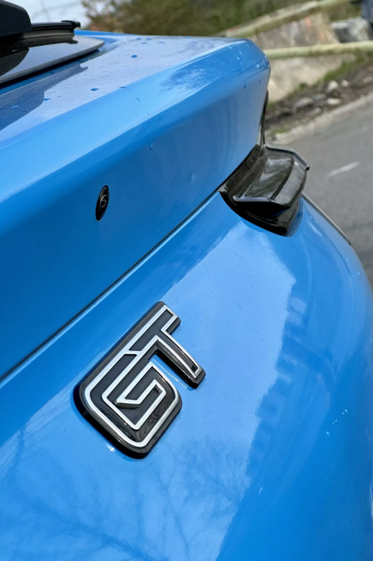 Zbliżenie tylnego odcinka niebieskiego Forda Mustang Mach-E z 2020 roku z widocznym metalowym emblematem GT, zakrzywionym bagażnikiem i aerodynamicznym tylnym spojlerem z czarną końcówką na delikatnie rozmytym tle.