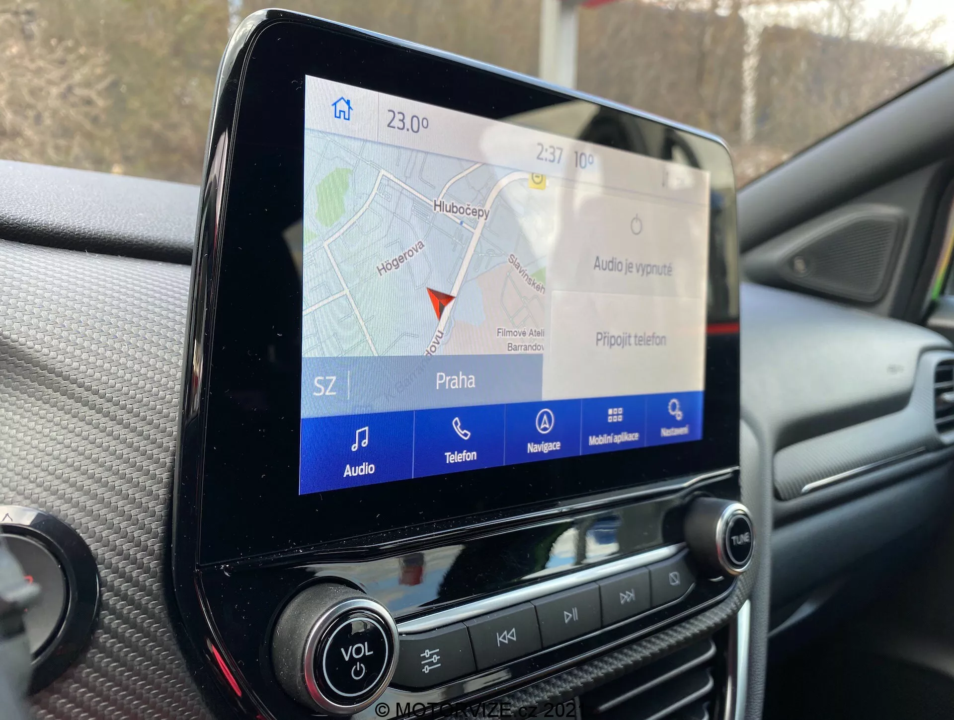 Zbliżenie na wyświetlacz dotykowy systemu infotainment wewnątrz Forda Puma 2020, przedstawiający mapę nawigacyjną GPS z oznaczeniem 'Praha', z ikonami funkcji audio, telefonu i nawigacji, otoczonymi przez fizyczne przyciski i pokrętła na desce rozdzielczej.