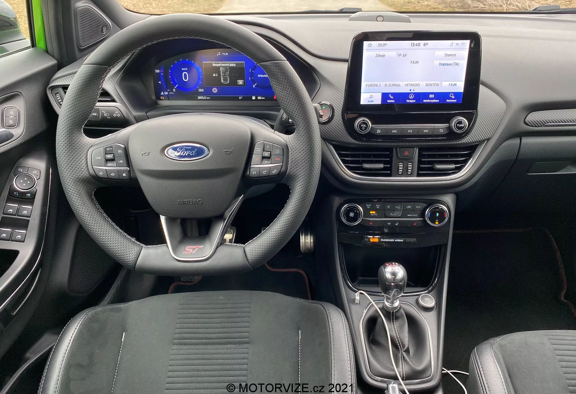 Widok wnętrza Ford Puma ST z 2020 roku z tyłu, podkreślający skórzaną kierownicę z logo Forda i przyciskami sterującymi, cyfrowy zestaw wskaźników z niebieskim podświetleniem, centralny ekran dotykowy systemu infotainment, nowoczesną konsolę środkową z kontrolą klimatyzacji, manualną dźwignię zmiany biegów z osłoną skórzaną i metalowymi akcentami, profilowane fotele sportowe oraz wysokiej jakości materiały, w tym skórę, tworzywa sztuczne i metalowe akcenty.