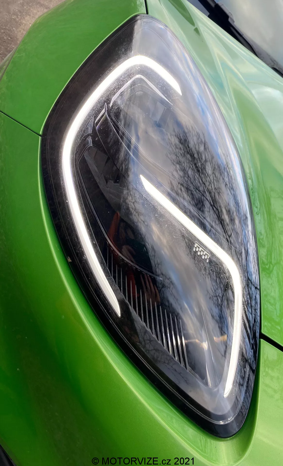 Zbliżenie lampy przedniej po lewej stronie (strona kierowcy) Forda Puma ST 2020 w żywym zielonym kolorze. Obraz ukazuje elegancki i kanciasty design reflektora pod kątem ukośnym, podkreślając jego nowoczesne elementy LED i błyszczącą, refleksyjną powłokę lakierniczą.
