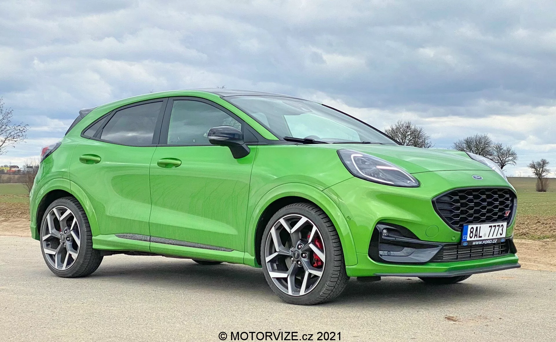 Jasnozielony hatchback Ford Puma ST 2020 sfotografowany pod kątem bocznym, ukazujący profil auta od przodu do tyłu. Niskokątny kadr podkreśla wyraźny grill o strukturze plastra miodu, sportowe reflektory, konturowaną maskę i duże felgi aluminiowe z czerwonymi zaciskami hamulcowymi. Elegancki, aerodynamiczny design obejmuje stopniowo opadającą linię dachu, widoczne linie charakteru i zarys tylnego spojlera.