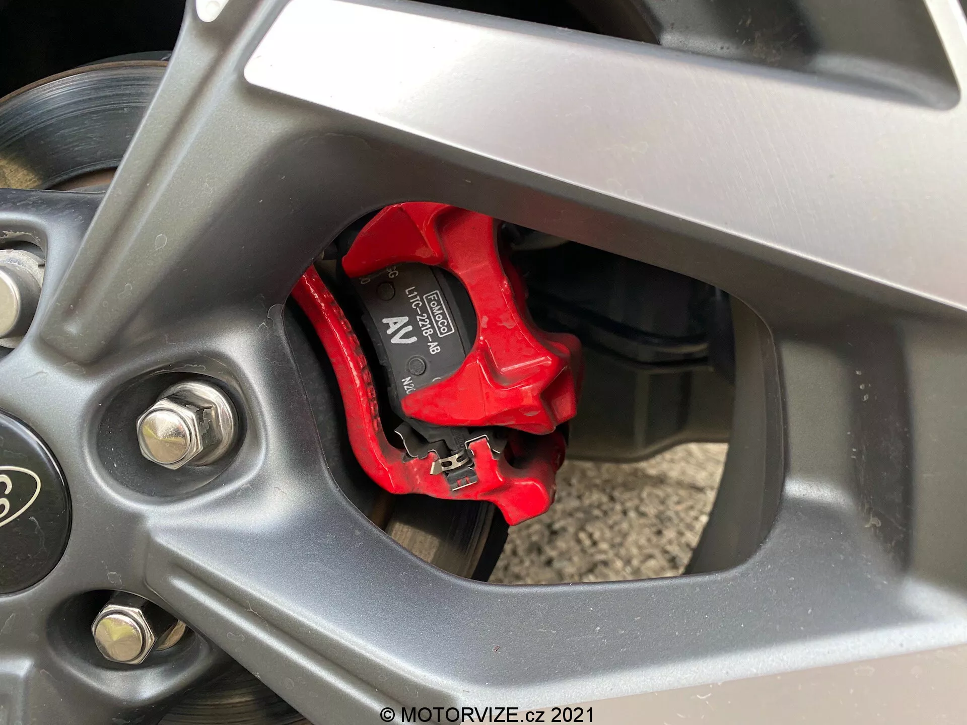 Szczegółowy zbliżenie na koło pojazdu Ford Puma ST 2020, skoncentrowane na wysokowydajnym czerwonym zacisku hamulca Brembo i wentylowanej tarczy hamulcowej. Koło o pięciu rozgałęzionych szprychach ma ciemne metaliczne wykończenie, z widocznymi srebrnymi śrubami mocującymi i częściowo widoczną ścianą boczną opony. Komponenty zawieszenia widoczne w tle.