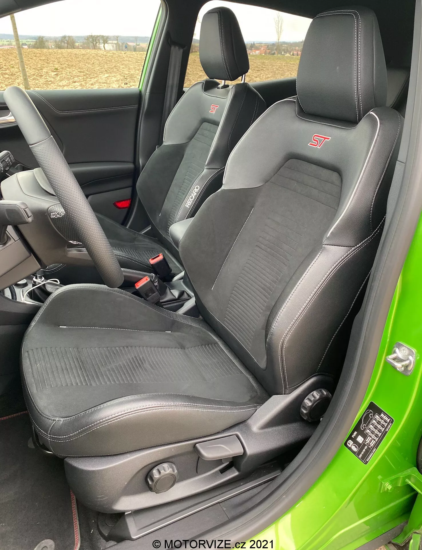 Zdjęcie wnętrza Ford Puma ST 2020, zrobione od otwartych drzwi po stronie kierowcy, skupiające się na sportowym fotelu kierowcy. Fotel ma zintegrowany zagłówek, wyraźne podparcie boczne i udowe oraz tapicerkę będącą połączeniem skóry i tkaniny z teksturowanymi sekcjami materiałowymi. Na górnej części fotela widoczne jest haftowane logo 'ST'. Widzoczne są dźwignie regulacji manualnej i pokrętło do regulacji podparcia lędźwiowego. W tle częściowo widoczna jest kierownica i pas bezpieczeństwa, a wnętrze jest oświetlone naturalnym światłem dziennym.