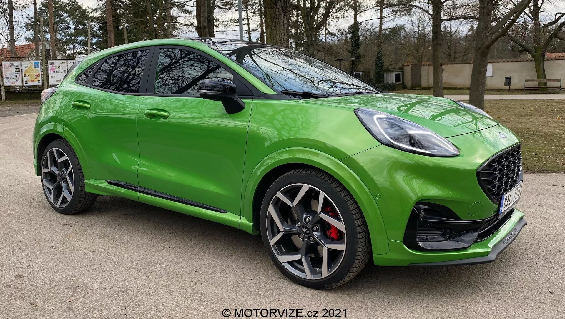 Jaskrawozielony Ford Puma ST 2020 sfotografowany z niskiego trzyczwartego kąta z przodu, podkreślający jego elegancki i sportowy design z grillem w kształcie plastra miodu, ostrymi reflektorami LED, ciemnymi wieloramiennymi felgami aluminiowymi, czerwonymi zaciskami hamulcowymi, wyrzeźbionym profilem bocznym, lusterkami z wbudowanymi kierunkowskazami, opadającą linią dachu, błyszczącym wykończeniem lakieru i dodatkowymi aerodynamicznymi detalami zewnętrznymi. Samochód jest zaparkowany na zewnątrz z rozmytymi drzewami i budynkiem w tle.