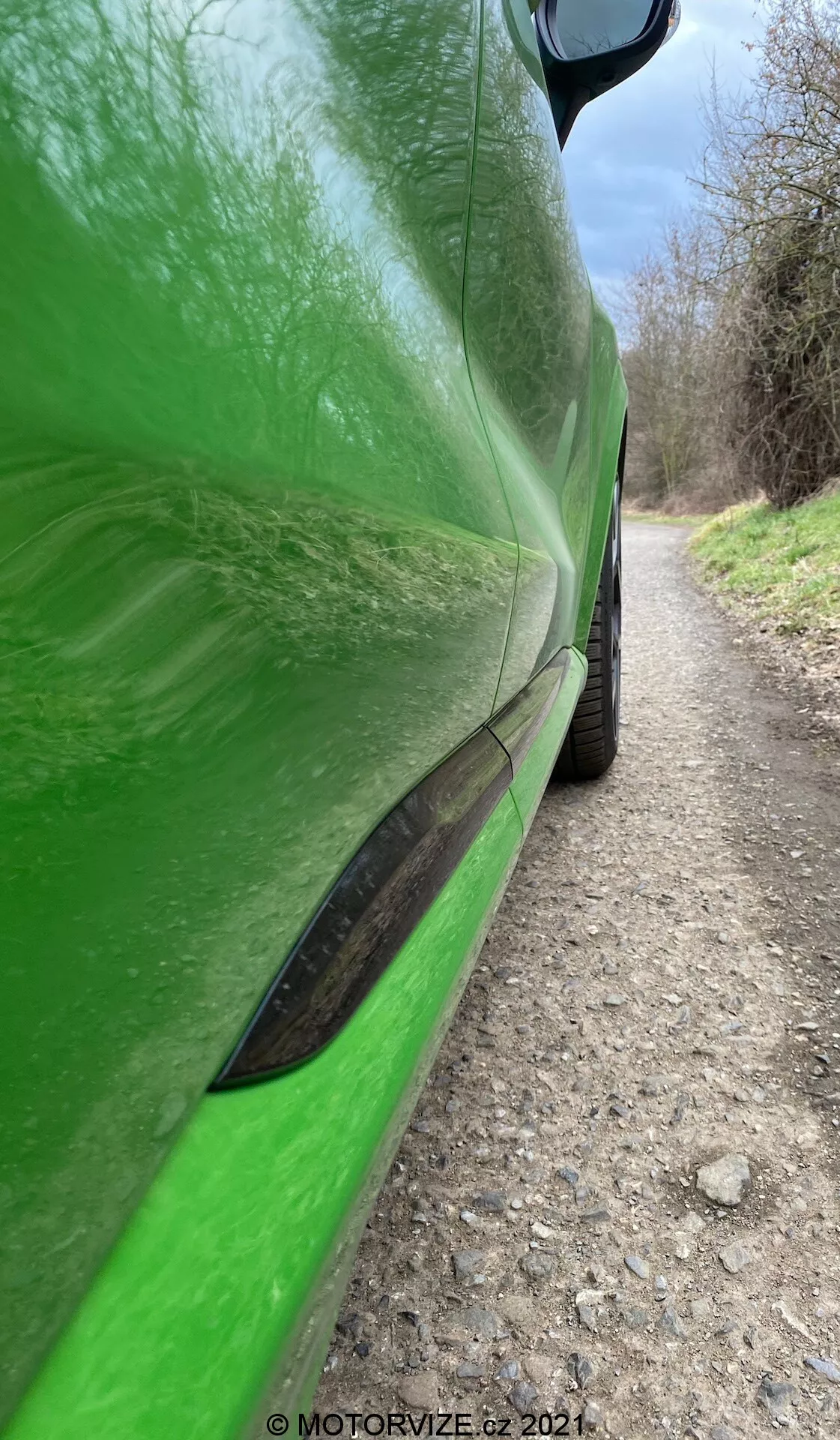 Zbliżenie boczne jasnozielonego samochodu Ford Puma 2020 (Puma-ST), pokazujące szczegóły od tylnego nadkola do lusterka bocznego, z naciskiem na obszar drzwi kierowcy. Wykończenie lakierem o wysokim połysku odbija naturalne otoczenie, w tym drzewa i ścieżkę. Charakterystyczne cechy to elegancka czarna listwa boczna, wyraźna linia karoserii na drzwiach i wąski wlot powietrza w dolnej części drzwi. Widoczne są górna część tylnego nadkola i opona, jak również czarna obudowa lusterka bocznego w prawym górnym rogu. Obraz podkreśla dynamiczne linie i kontury samochodu.