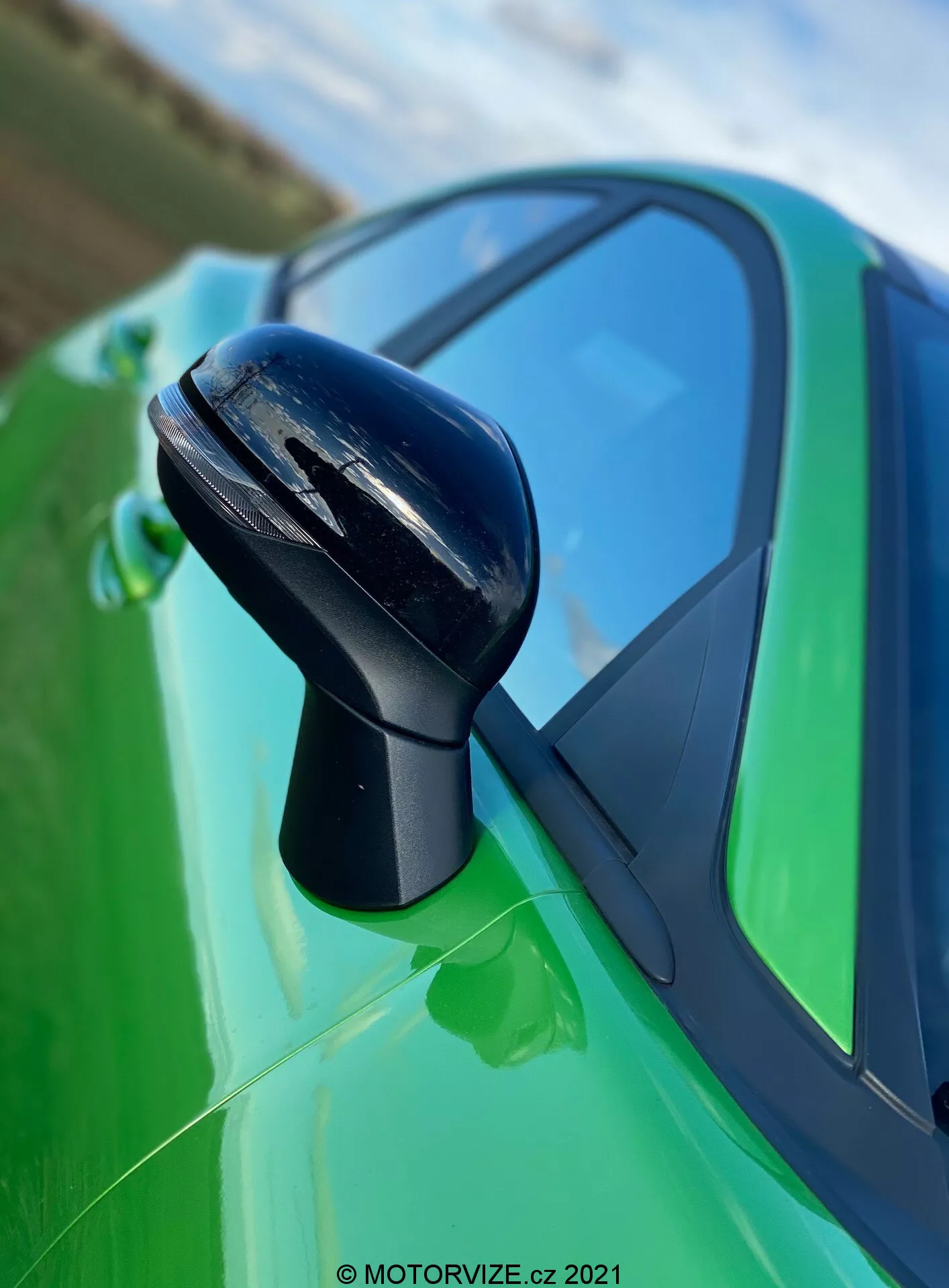 Zbliżenie na lusterko boczne po stronie kierowcy w Fordzie Puma ST 2020, prezentujące elegancki czarny aerodynamiczny design z możliwymi zaawansowanymi funkcjami, takimi jak wskaźniki kierunkowskazów lub zintegrowany system monitorowania martwego pola. Powierzchnia odbijająca lusterka i czarna ramka pasują do jego obudowy, przylegającej do lśniącej limonkowej karoserii z błyszczącym wykończeniem, z widocznym przyciemnionym tylnym oknem i sportowym spojlerem. Tło stanowi niebieskie niebo z rozmazanym naturalnym otoczeniem.