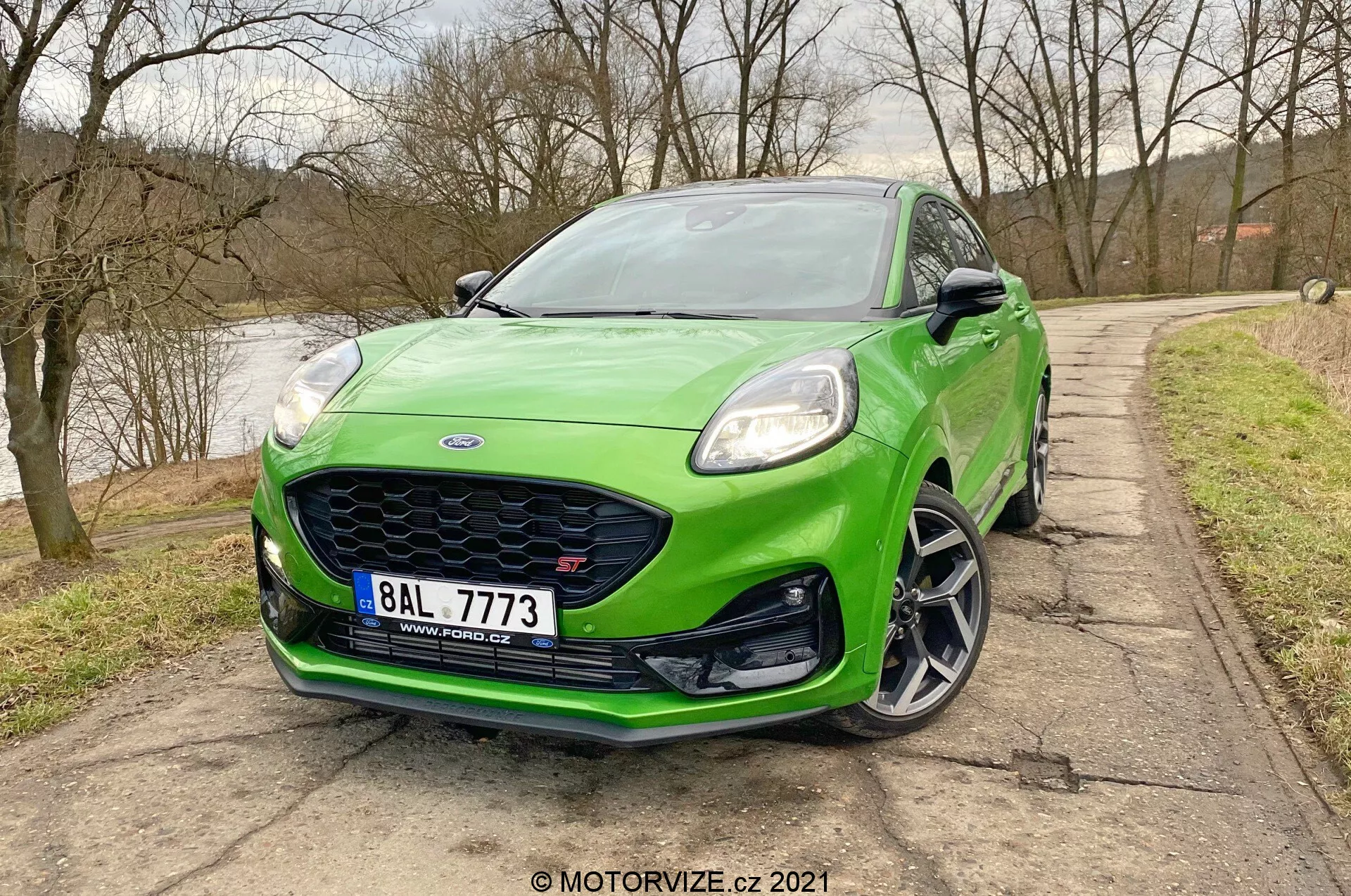 Widok z przodu w trzech czwartych na jaskrawozielony samochód hatchback Ford Puma ST 2020, ukazujący przednie elementy designu, w tym lewy reflektor, przedni zderzak, grill i kontur lewego błotnika. Widoczny jest również elegancki kaptur i przednia szyba. Obraz jest uchwycony pod delikatnym kątem w dół, podkreślając agresywny przedni zderzak z wlotami powietrza i splitterem, eleganckie reflektory LED, wieloramienne koła z niskoprofilowymi oponami oraz zintegrowany kierunkowskaz w lusterku bocznym. Zbliżenie podkreśla sportowy i nowoczesny wygląd pojazdu.