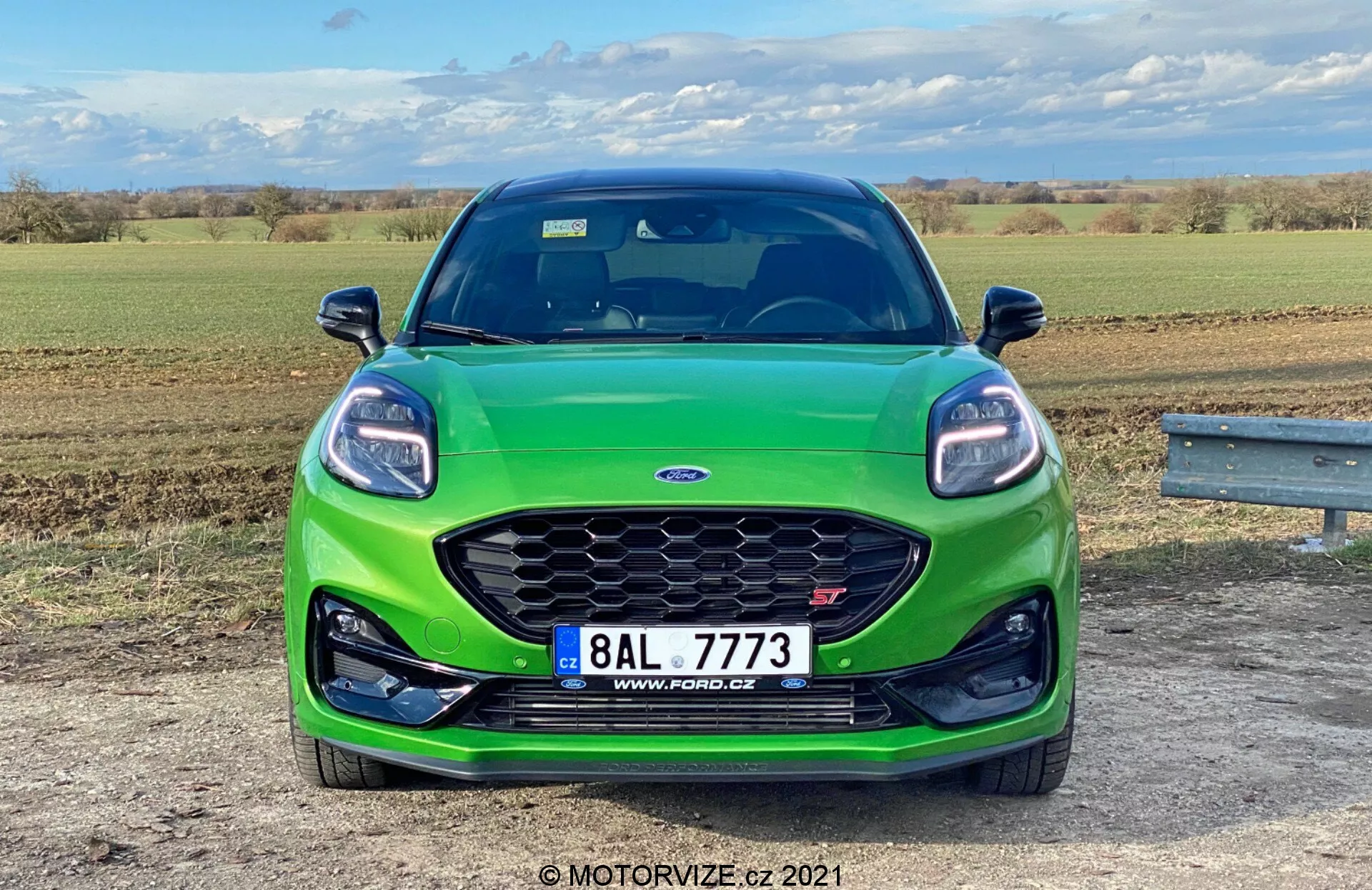 Widok z przodu zielonego Forda Puma ST 2020 zaparkowanego na otwartym terenie naturalnym w ciągu dnia, ukazujący sportowy przedni grill z siatkowym wzorem, wydłużone reflektory LED, centralną tablicę rejestracyjną i czarną sekcję przedniego zderzaka, prawdopodobnie wyposażoną w zintegrowane czujniki.