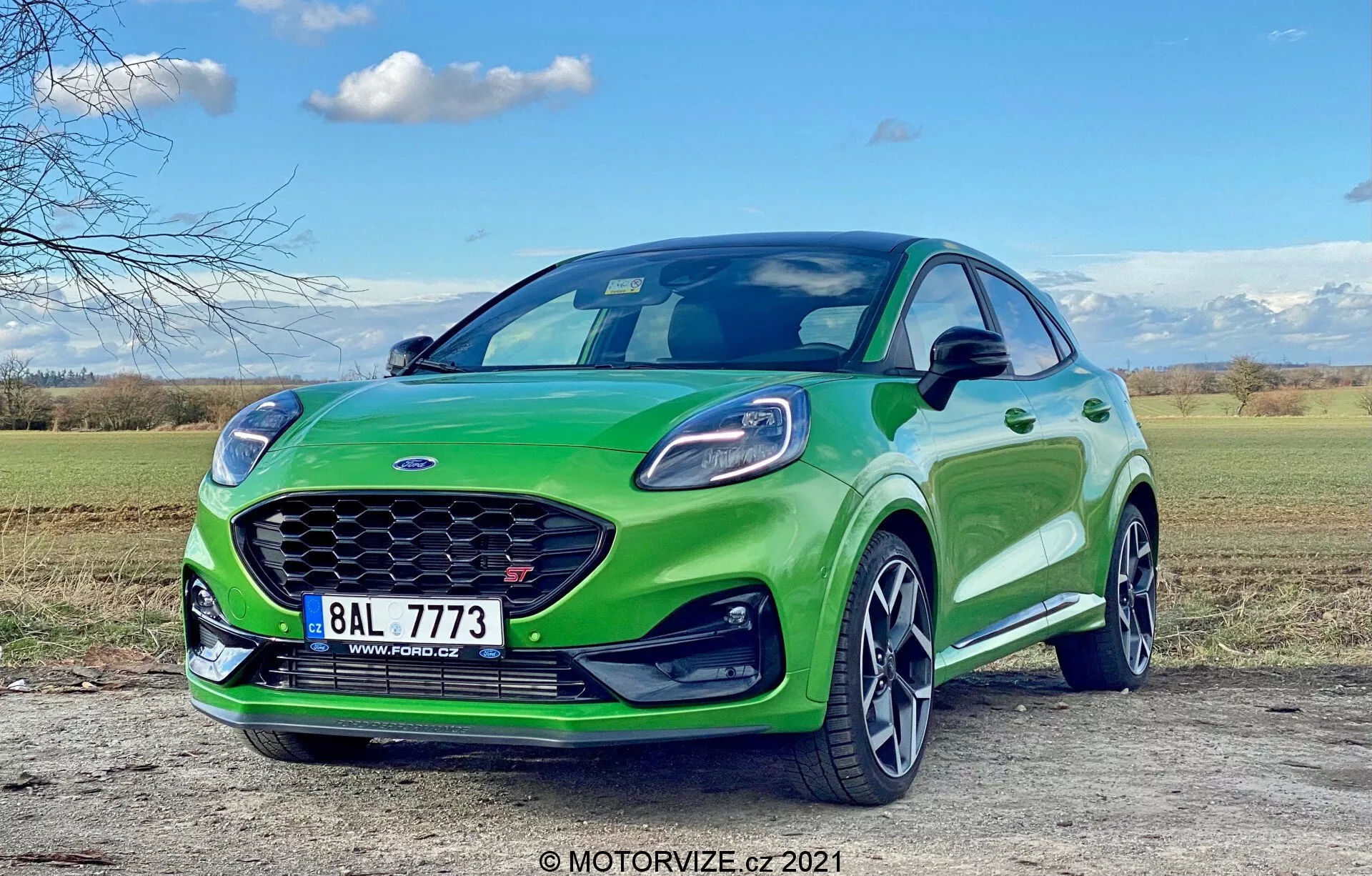 Widok z niskiej perspektywy na Forda Puma ST 2020, który pokazuje przednią stronę kierowcy z dziennymi światłami LED w konstrukcji reflektora, dużym przednim grillem, aerodynamicznymi konturami maski, felgami aluminiowymi i lusterkami bocznymi z zintegrowanymi kierunkowskazami. Żywo zielony samochód jest zaparkowany na zewnątrz za dnia, kontrastując z czystym niebem i naturalnym krajobrazem.