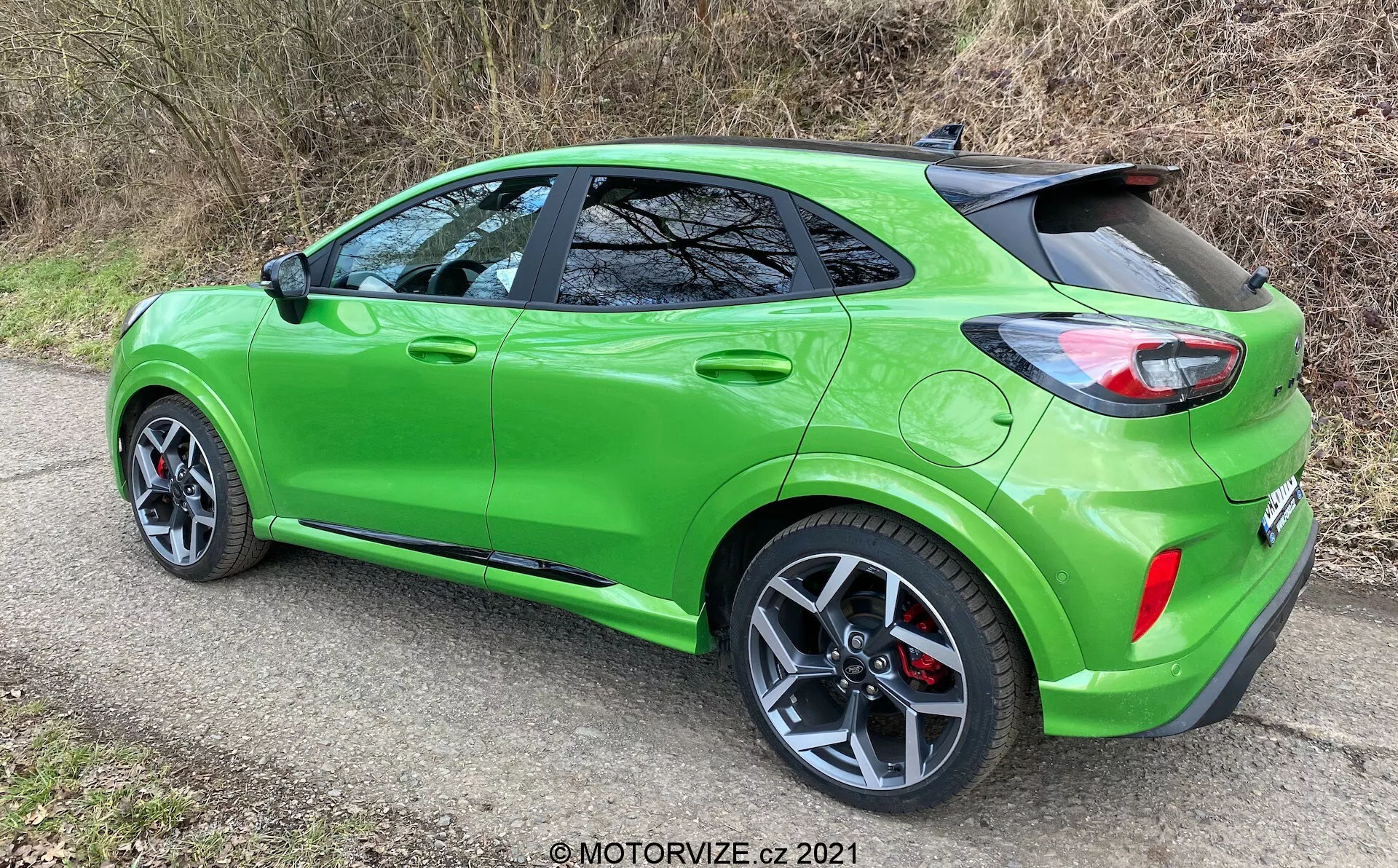Jaskrawo zielony kompaktowy crossover SUV Ford Puma ST 2020 widziany z tyłu pod kątem trzech czwartych, pokazujący wieloramienne felgi aluminiowe, opony o niskim profilu, opadającą linię dachu, nowoczesne tylne światła, spojler dachowy, wyraźne nadkola, żywo zielony lakier, przyciemniane tylne szyby, podwójne końcówki wydechu oraz nieidentyfikowalne znaki w warunkach dziennych z naturalnym otoczeniem.