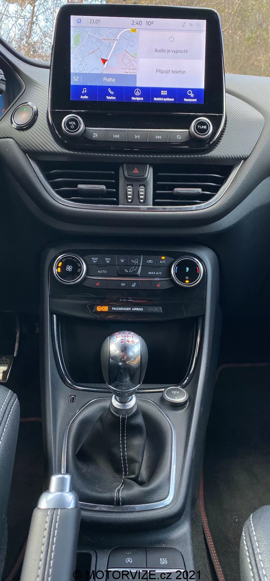 Zbliżony pionowy widok środkowej konsoli Ford Puma 2020, przedstawiający ekran dotykowy systemu infotainment wyświetlający mapę, sterowanie klimatyzacją i manualną dźwignię zmiany biegów z dodatkowymi kontrolkami trybów jazdy. Konsola zawiera mieszankę miękkiego plastiku i skóry z kontrastowymi szwami, podkreślając jakość wnętrza i ergonomię samochodu.