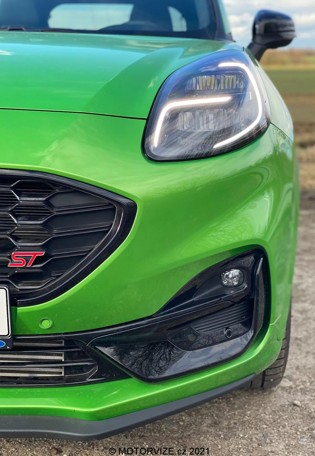 Zbliżenie na przednią lewą część jasnozielonego Forda Puma ST z 2020 roku, pokazujące lewą lampę LED z nowoczesnym kanciastym wzorem i światłami do jazdy dziennej LED, kratkę o wzorze plastra miodu z czerwonym znakiem 'ST' oraz kanciaste czarne obudowy na przednim zderzaku, prawdopodobnie do przednich świateł przeciwmgielnych lub czujników. Niski kąt podkreśla sportową postawę, wyróżniając cechy związane z osiągami i zaawansowane technologicznie elementy designu.