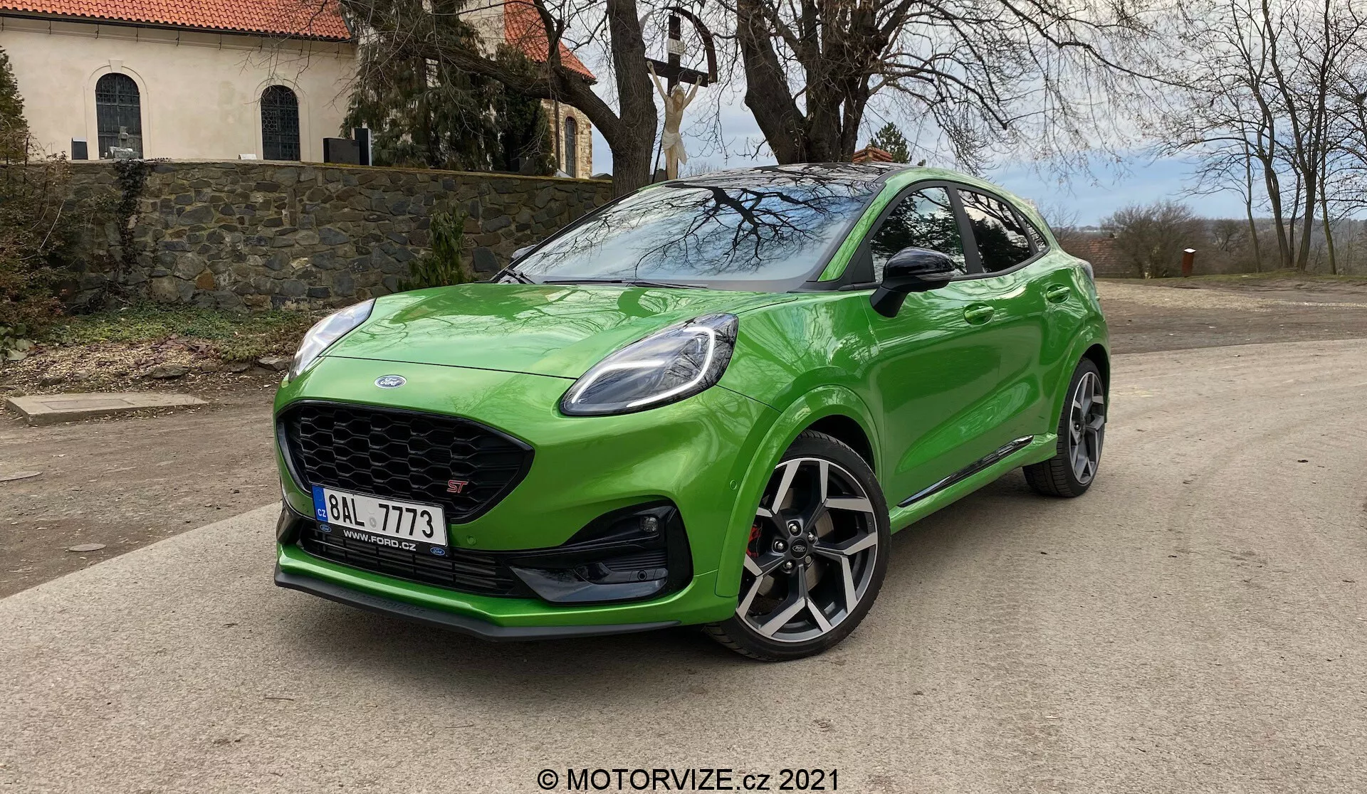 Obraz przedstawia jasnozielony hatchback Ford Puma ST 2020 zaparkowany na utwardzonej powierzchni widoczny pod kątem trzech czwartych od przodu. Samochód ma dużą kratkę o strukturze plastra miodu, aerodynamicznie stylizowany przedni zderzak z wlotami powietrza, ostre reflektory LED lub HID z dziennymi światłami, oraz maskę z dynamicznymi liniami. Profil po stronie kierowcy podkreśla duże wieloszprychowe felgi aluminiowe, progi boczne i kompaktową sportową sylwetkę z opadającą linią dachu. Szyba przednia jest pochylona dla aerodynamicznego profilu z lekko przyciemnionymi szybami. Błyszczące zielone wykończenie samochodu i naturalne światło dzienne z miękkimi cieniami pod samochodem dodają mu nienagannego wyglądu. Widoczne są loga na kratce i tablica rejestracyjna z przodu, osadzone na tle naturalnym z drzewami.