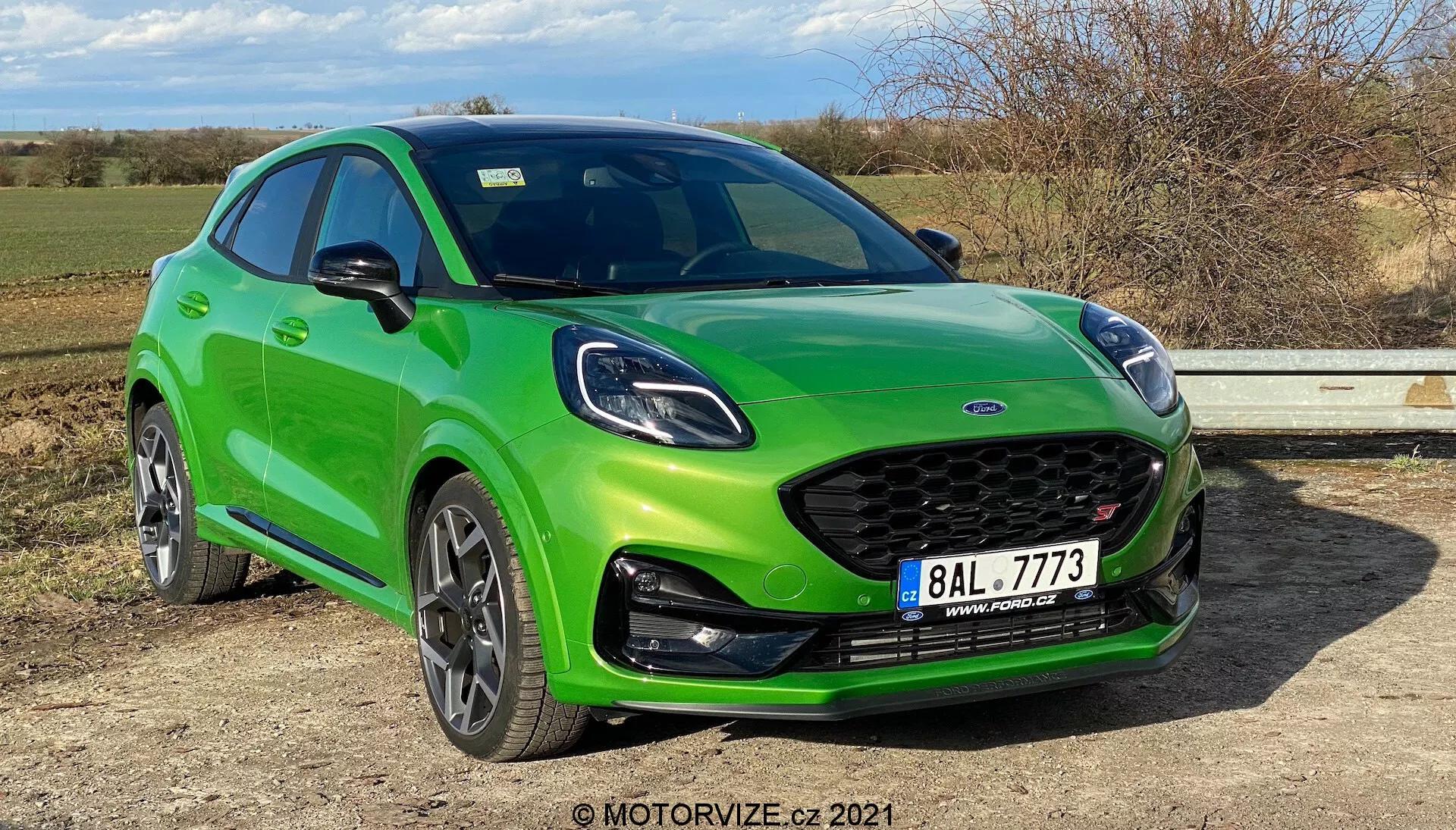 Żywiołowo zielony Ford Puma ST 2020 pokazany z trzech czwartych z przodu z lewej strony, podkreślający boczny profil i przednią część pojazdu. Charakterystyczna osłona chłodnicy z emblematem producenta na środku, sportowy zderzak z wbudowanymi światłami przeciwmgielnymi i spojlerem dolnym. Maskę charakteryzują sportowe kontury, wąskie reflektory z dziennymi światłami LED. Dynamiczny boczny profil hatchbacka ma opadającą linię dachu, lusterka boczne w kolorze nadwozia, czarne listwy ochronne wzdłuż dolnej części drzwi i nadkoli, wyraźne felgi aluminiowe, mogły być wyposażone w systemy monitorowania ciśnienia w oponach, a także widoczne tarczowe hamulce. Samochód zaparkowany jest na płaskiej powierzchni, z błyszczącym wykończeniem lakierniczym, ogólnie podkreślającym jego sportowy design, technologię oświetlenia i funkcje wydajnościowe w naturalnym świetle dziennym.