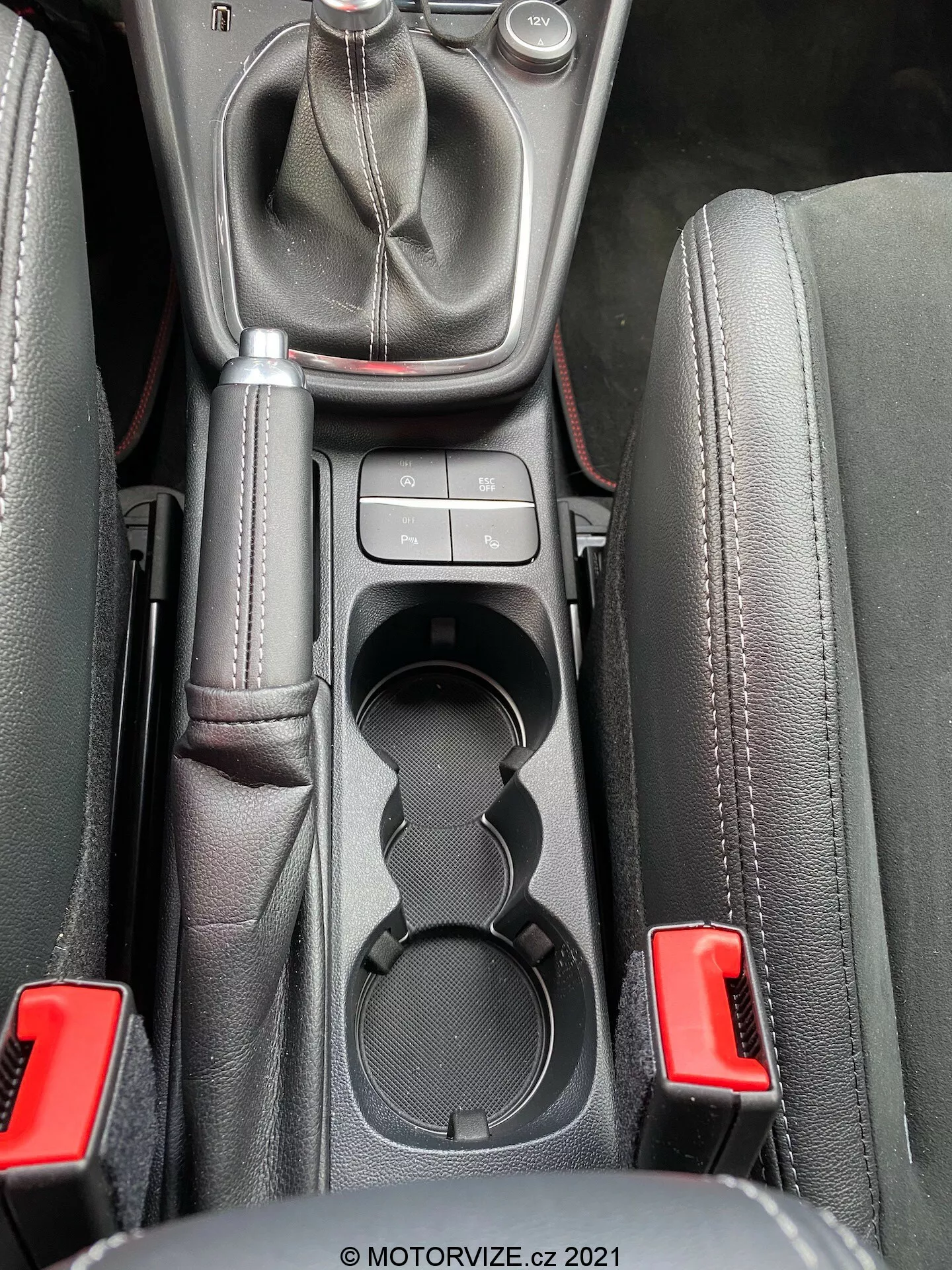 Zbliżenie wnętrza Ford Puma ST 2020, skupione na obszarze konsoli środkowej widzianym z wysokości oczu, ukazujące manualny lewarek zmiany biegów z osłoną skórzaną i metalową górką, otoczony przez elementy sterujące konsoli centralnej, podwójne uchwyty na kubki, wyściełany podłokietnik środkowy i klamry pasów bezpieczeństwa w naturalnym świetle.