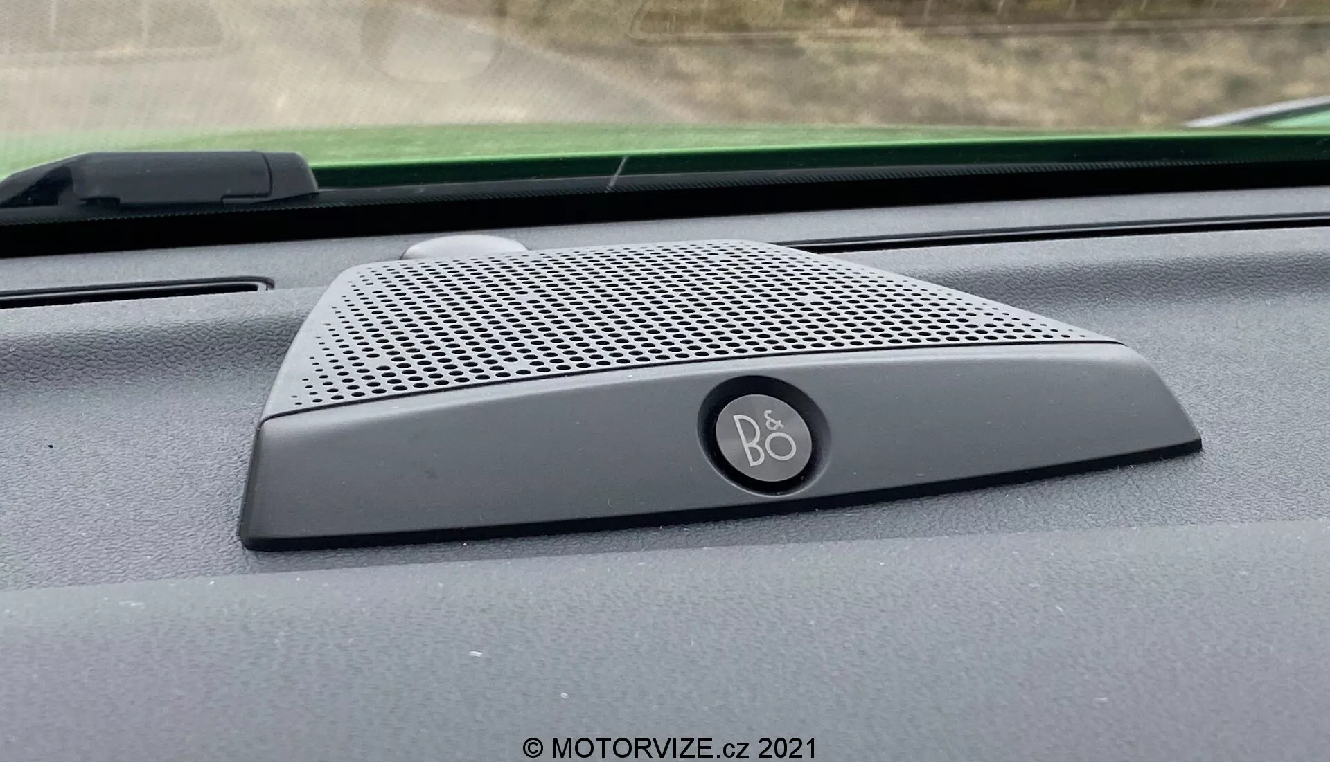 Zbliżenie wnętrza głośnika deski rozdzielczej Ford Puma ST 2020 z logo B&O (Bang & Olufsen), charakteryzujący się eleganckim designem z drobno perforowaną kratką i zaokrąglonymi krawędziami, umieszczony u podstawy przedniej szyby, podkreślający integrację systemu audio premium.