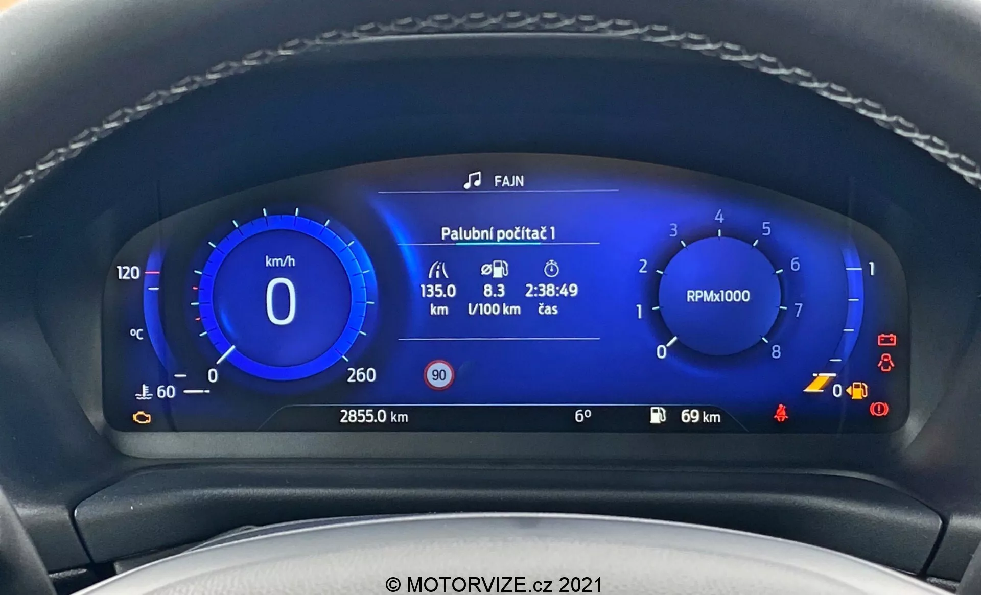 Zbliżenie nowoczesnego cyfrowego wyświetlacza deski rozdzielczej Ford Puma ST 2020 z perspektywy kierowcy. Obraz podkreśla żywy niebieski cyfrowy zestaw wskaźników obramowany krawędzią kierownicy. Tablica rozdzielcza pokazuje prędkościomierz, obrotomierz, dane dotyczące zużycia paliwa, całkowity przebieg, temperaturę zewnętrzną oraz różne symbole ostrzegawcze, ilustrując zaawansowane technologie motoryzacyjne.