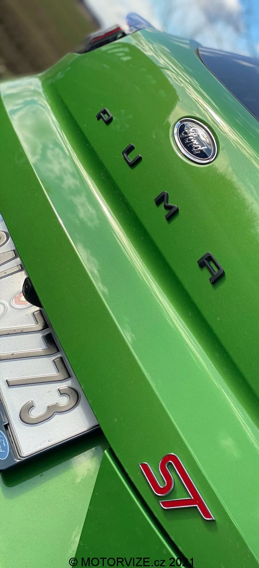 Zbliżenie tylnego widoku z niskiego kąta jasnozielonego Forda Puma 2020. Widoczny jest poziomy tylny spojler z centralnym, okrągłym emblematem, chromowane litery 'PUMA' oraz sportowy czerwony znak 'ST', wskazując na usportowioną wersję modelu. Karoseria odbija jasne warunki oświetleniowe na zewnątrz.