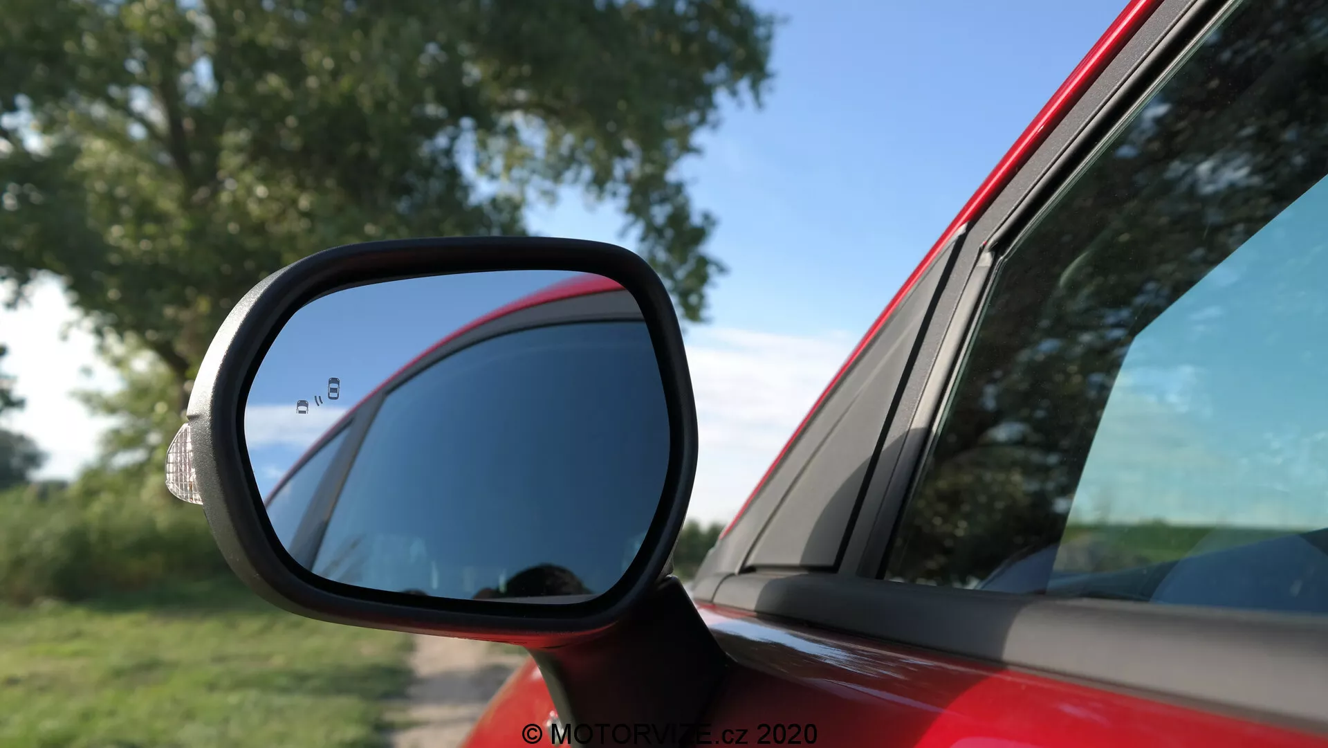 Zbliżenie lusterka bocznego w czerwonym Fordzie Puma 2020, skoncentrowane na aerodynamicznej, czarnej obudowie z tradycyjnym płaskim lustrem i sekcją martwego pola. Zdjęcie pokazuje drzwi kierowcy i tylny panel w świetle dziennym, podkreślając krzywizny i linie projektowe samochodu. Scena sugeruje, że pojazd jest zaparkowany na otwartym terenie wśród naturalnego otoczenia.
