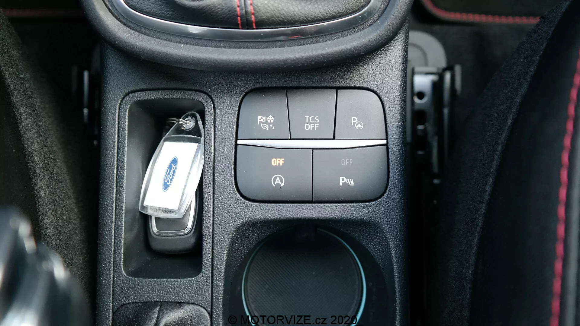 Zbliżenie na konsolę centralną Ford Puma 2020 z perspektywy kierowcy, pokazujące zestaw zaawansowanych technologicznie przycisków i przełączników do kontrolowania funkcji samochodu. Konsola zawiera przycisk czujników parkowania, przycisk wyłączania systemu kontroli trakcji (TCS), dwa przełączniki zasilania i miejsce na gniazdo 12V. Klucz do samochodu wisi nad środkową częścią konsoli, która jest zaakcentowana teksturowaną czarną ramką z tworzywa sztucznego i czerwonymi szwami, dodającymi luksusowego charakteru.