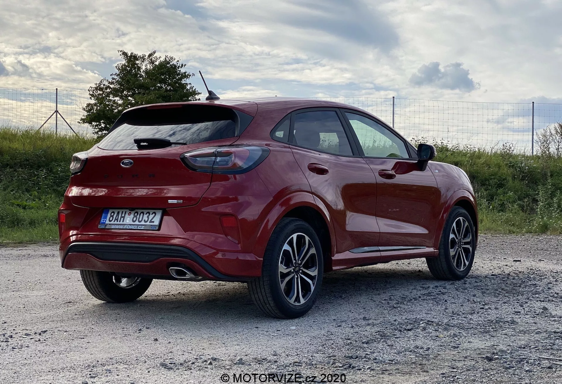 Na tym zdjęciu widać nowoczesny crossover SUV Ford Puma 2020 zaparkowany na żwirowej powierzchni. Zdjęcie wykonane z ujęcia z trzech czwartych tylnej części uwypukla boczny profil i tylną część, pokazując zakrzywioną klapę bagażnika z zintegrowanym spojlerem tylnym, wyeksponowanym logo, tylną wycieraczkę oraz poziome światła tylne. Metaliczny czerwony lakier lśni, ukazując połyskliwe wykończenie uzupełnione obręczami z wieloszprychowym wzorem. Karoseria pojazdu ma charakterystyczne linie i krzywizny zwiększające aerodynamikę, z rosnącą linią boczną i widocznymi relingami dachowymi. Oświetlenie późnym popołudniem lub wczesnym wieczorem podkreśla kolor i design samochodu. Potencjalne funkcje technologiczne mogą obejmować zaawansowane systemy wspomagania kierowcy (ADAS).