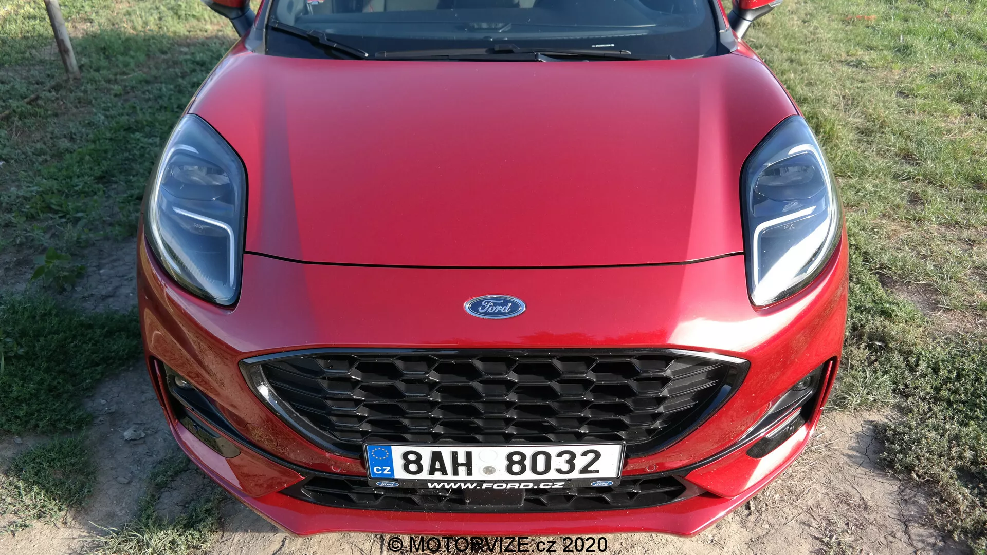 Widok z przodu czerwonego Forda Puma 2020 uchwycony z lekko podwyższonego kąta, featuring chromowaną kratkę w kształcie plastra miodu z logo Ford, stylowe reflektory z możliwymi elementami LED, gładko konturowaną maskę oraz tablicę rejestracyjną w stylu europejskim sugerującą, że zdjęcie zostało zrobione w Czechach.