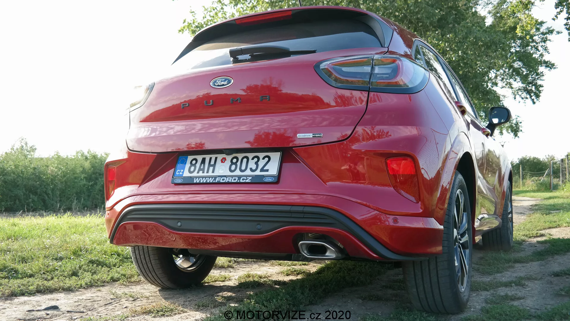 Zdjęcie czerwonego Forda Puma 2020 sfotografowanego pod kątem trzy czwarte od tyłu, ukazujące jego nowoczesny design hatchbacka/SUV-a. Pokrywa bagażnika ma zintegrowany spojler, rozległe poziome tylne światła LED i widoczne oznaczenia. Tylny zderzak zawiera zintegrowane reflektory i chromowaną końcówkę wydechu. Widoczna jest część tylnego koła z wieloramiennym wzorem. Samochód zaparkowany jest na świeżym powietrzu w naturalnym otoczeniu, co wskazuje na obszar podmiejski lub wiejski.