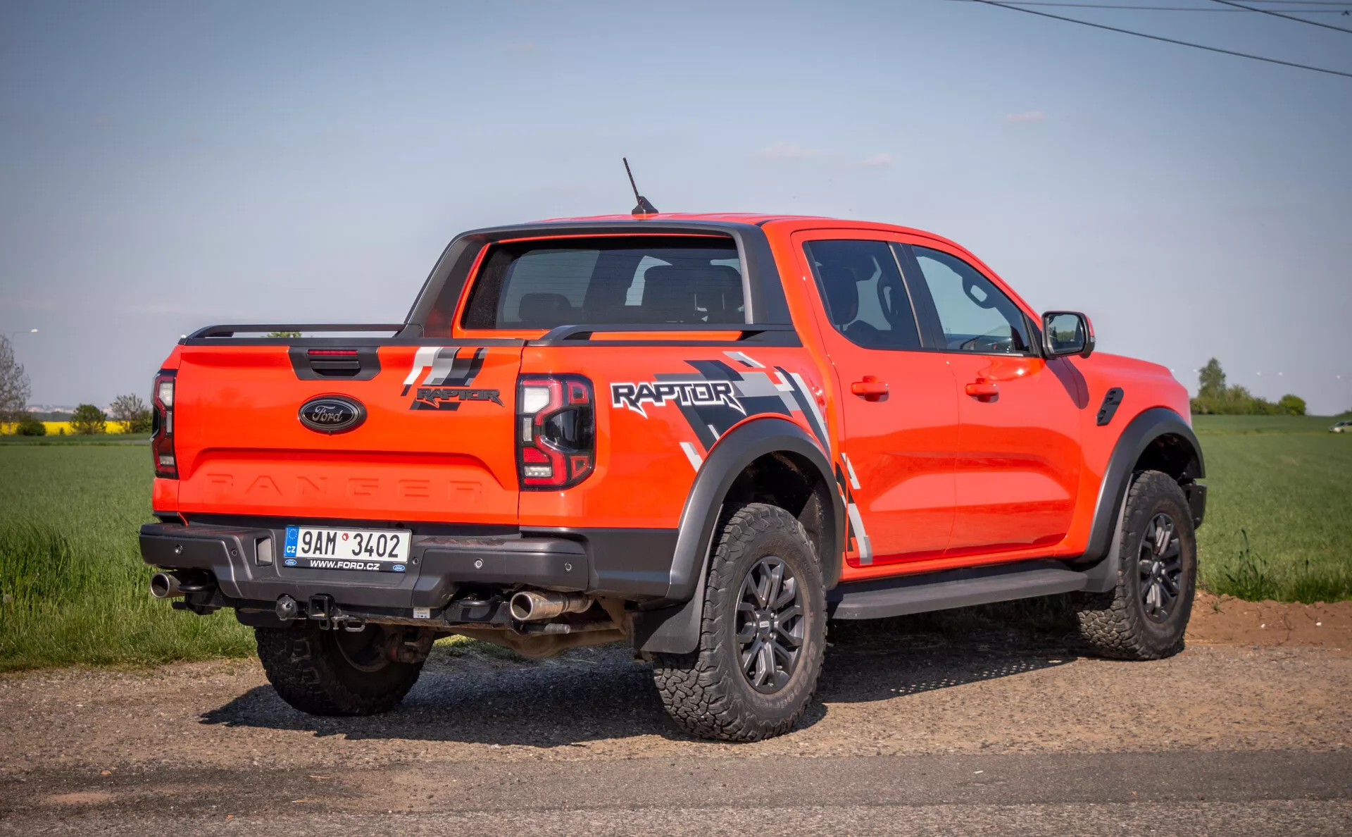 Jaskrawo pomarańczowy pickup Ford Raptor z 2022 roku w perspektywie boczno-tylnej, na zewnątrz na polu, prezentujący swoje zdolności terenowe z wysokim prześwitem, kabiną załogową, dużymi czarnymi osłonami nadkoli, czarnymi kołami o wieloramiennej konstrukcji z oponami terenowymi, oraz oznaczeniem 'RAPTOR' na klapie bagażnika.