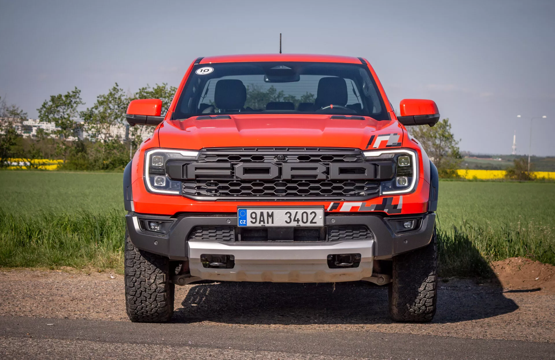 Przedni widok czerwonego pickupa Ford Ranger 2022 na asfalcie z tłem czystego nieba, przedstawiający grill, reflektory LED, przedni zderzak z hakami holowniczymi i płytą ochronną, oraz logo Forda