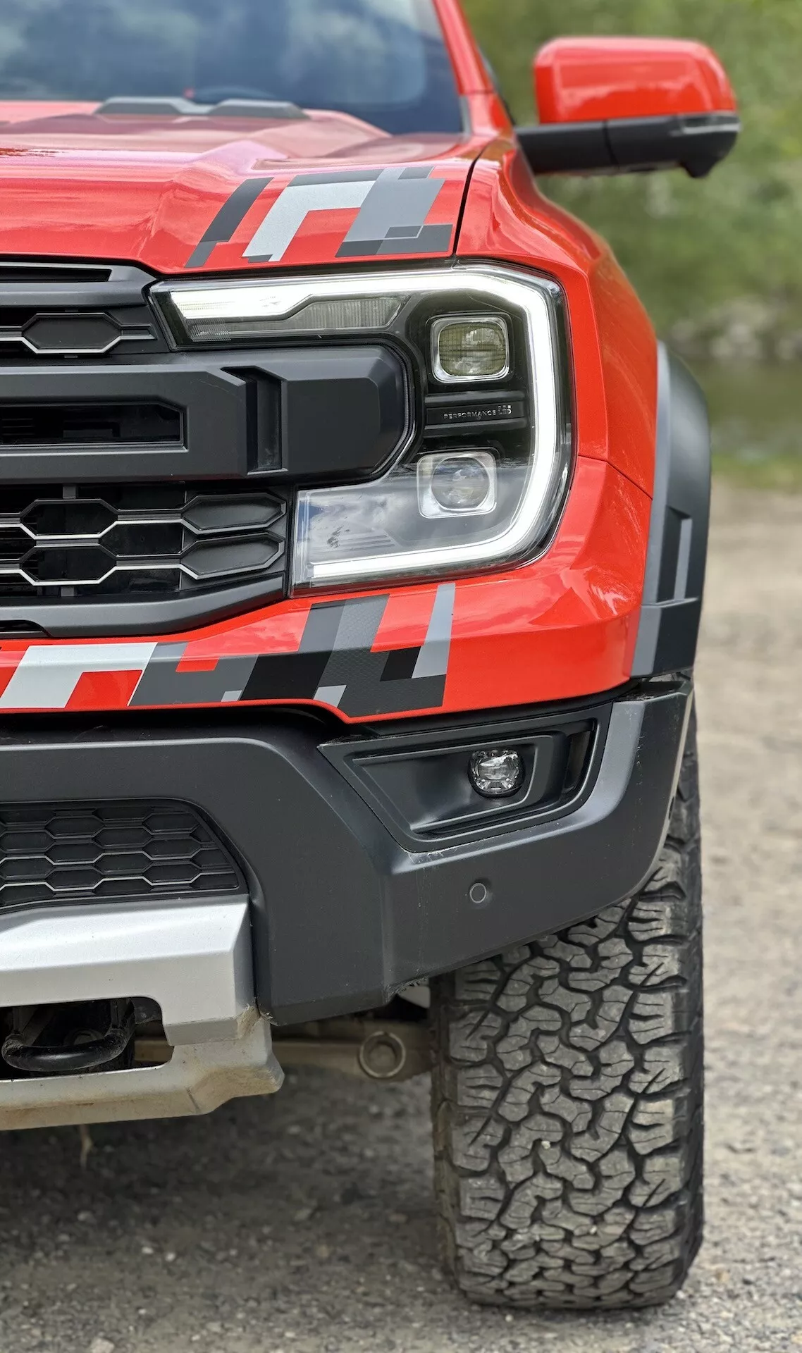 Zbliżony widok na czerwony pickup Ford Ranger 2022, prezentujący reflektor LED, solidną kratę, muskularny przedni błotnik z naklejką oraz bieżnik opony terenowej z niskiej perspektywy, aby podkreślić wytrzymałość i zaawansowaną technologię