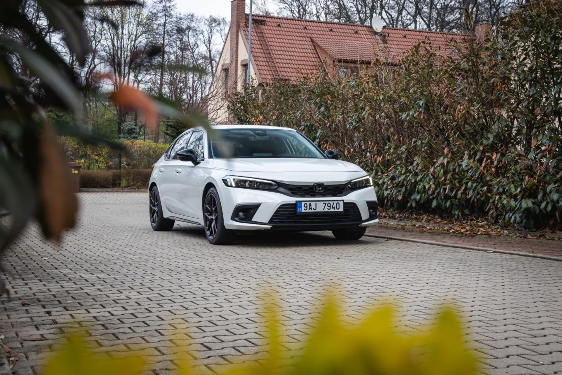 Nowoczesny sedan Honda Civic 2022 zaparkowany na bruku z zielenią i budynkiem w tle, prezentujący przedni grill, reflektory LED i elegancką aerodynamiczną konstrukcję