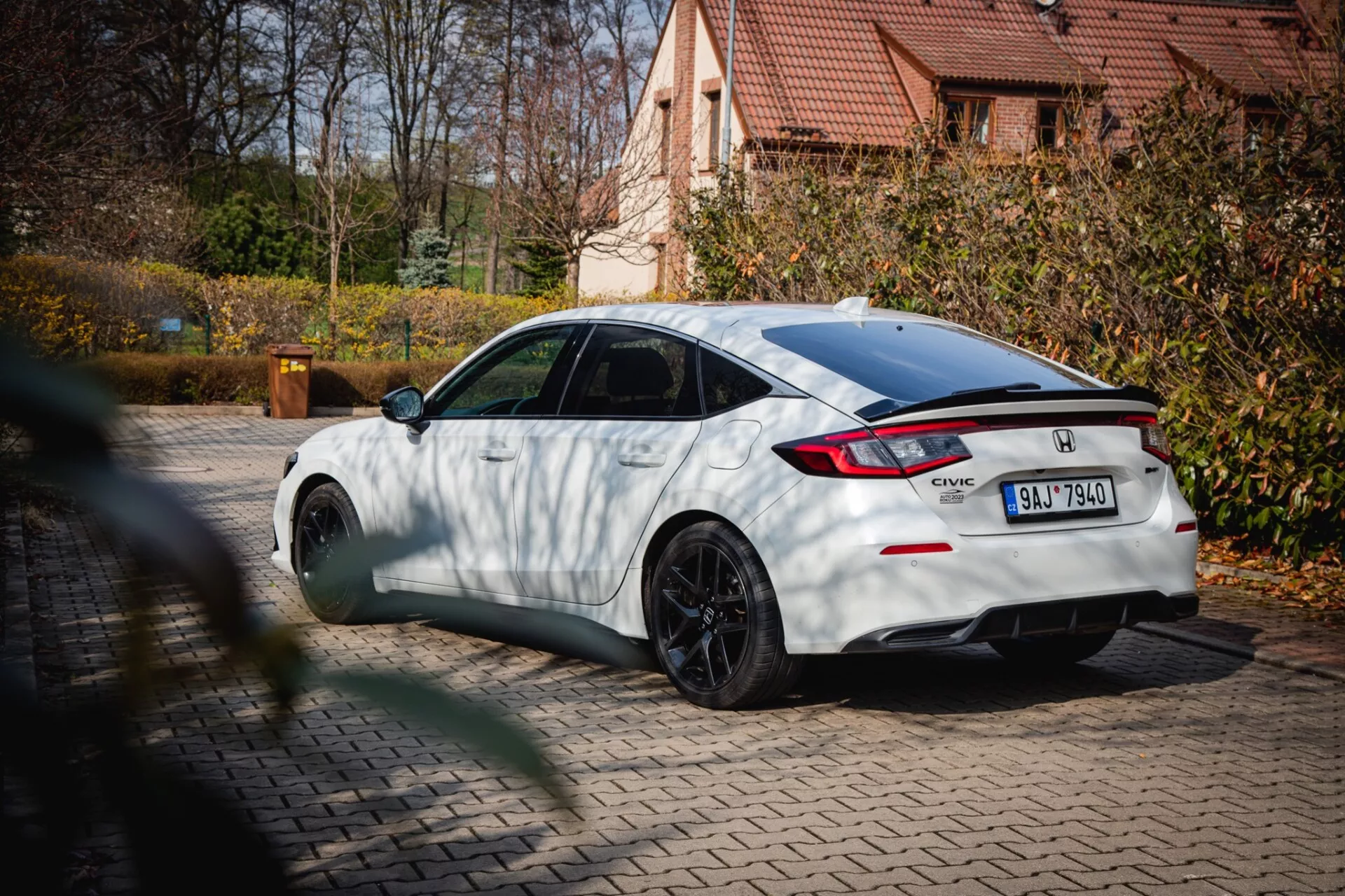 biały Honda Civic z 2022 roku z czarnymi kołami o wielu szprychach i tylnym spoilerem, zaparkowany na bruku z jesiennym liśćmi i mieszkaniowym domem w tle