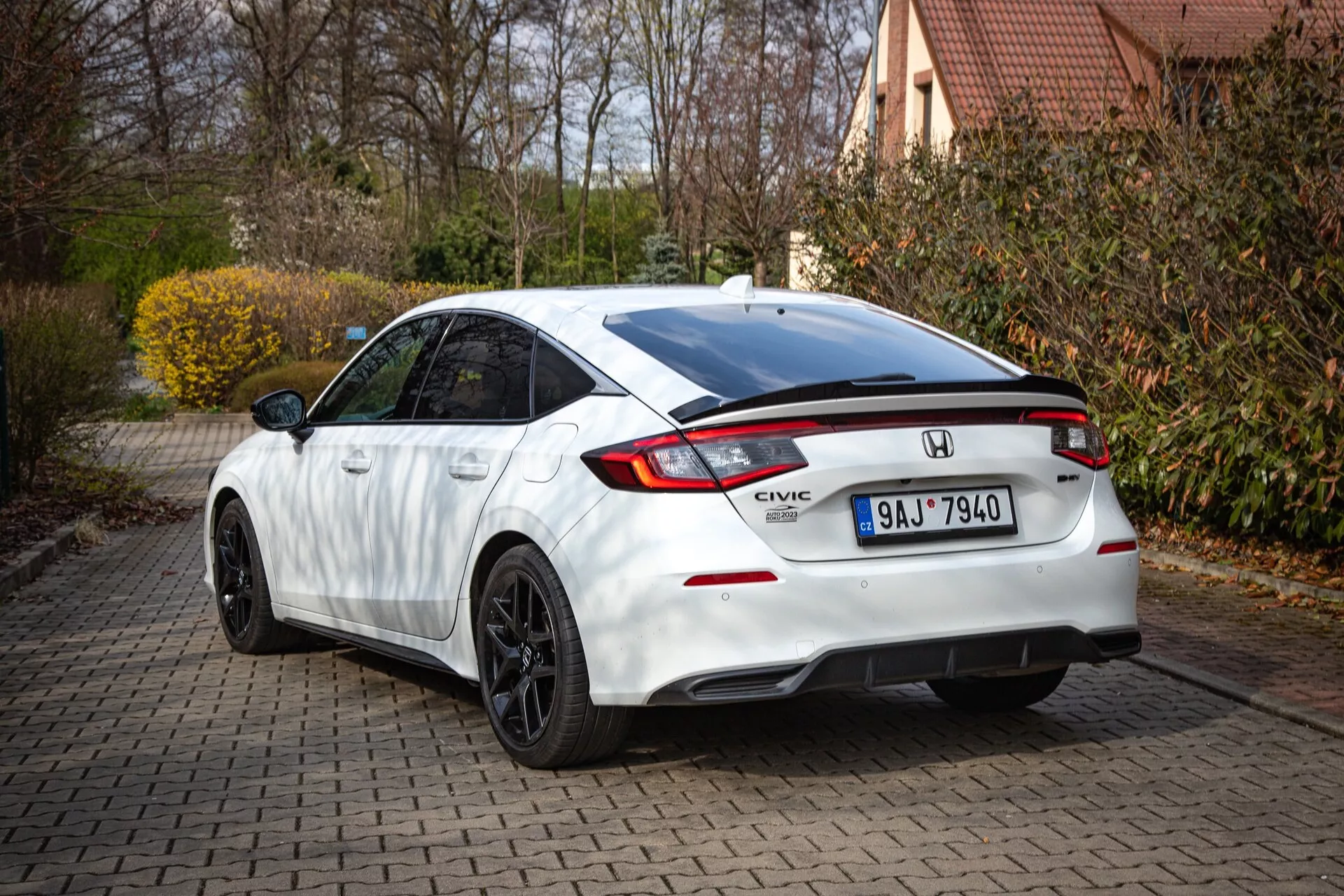Biały Honda Civic 2022 pokazujący tylny i częściowo boczny profil na ceglanej drodze dojazdowej, ze spadającą linią dachu, zintegrowanym spoilerm, światłem hamowania LED, charakterystycznymi światłami tylnymi i sportowym zderzakiem z dyfuzorem