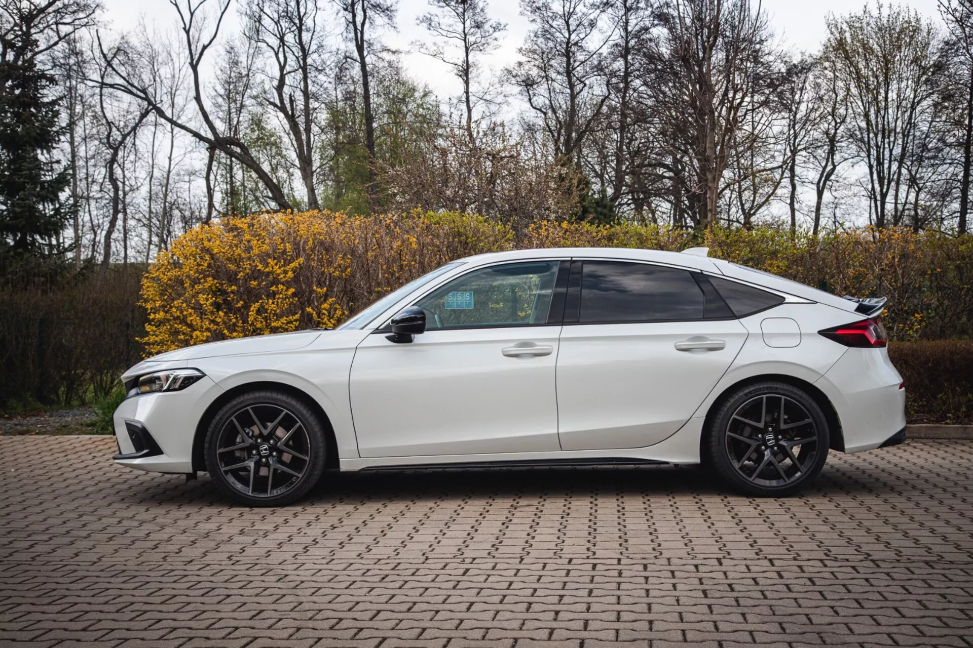 Biały czterodrzwiowy sedan Honda Civic z 2022 roku zaparkowany na brukowanej powierzchni z klocków, pokazujący profil boczny o sportowym wzornictwie i nowoczesnych reflektorach LED, uchwycony z lekko podniesionego kąta, aby podkreślić pełny widok boczny, w tym przyciemniane szyby, sportowe koła ze stopu i technologiczne funkcje, takie jak zintegrowane kierunkowskazy w lusterkach bocznych.