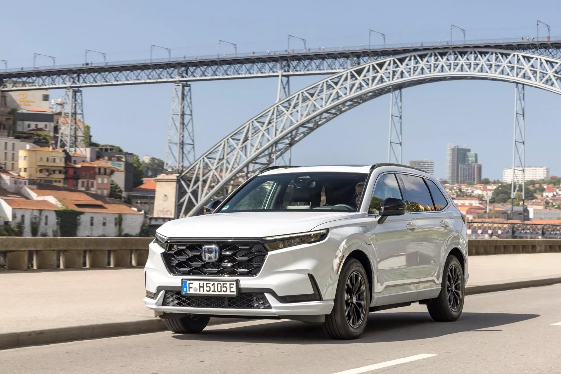 Honda CR-V 6. generacji 2023 jadąca po moście z reflektorami LED, dużym grillem z czujnikami bezpieczeństwa, aerodynamiczną konstrukcją, felgami aluminiowymi i kierunkowskazami zintegrowanymi z lusterkami bocznymi, w miejskim otoczeniu.