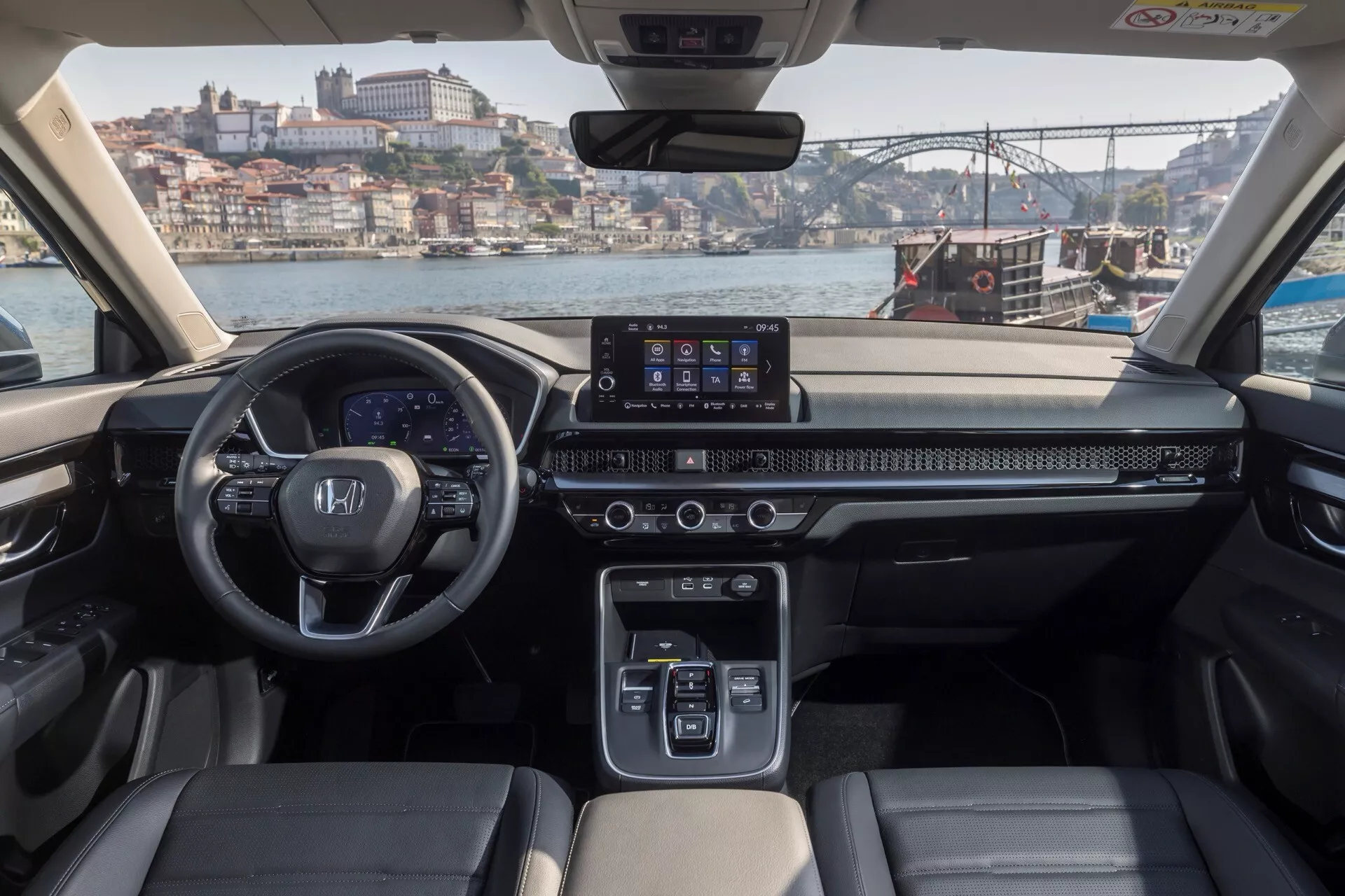 Wnętrze Hondy CR-V szóstej generacji 2023 prezentujące deskę rozdzielczą, cyfrowy zespół wskaźników, 10-calowy ekran dotykowy systemu infotainment, sterowanie klimatyzacją, selektor biegów z przyciskami, kierownicę z zintegrowanymi sterownikami, dwukolorowe skórzane siedzenia i panoramiczny widok na zewnątrz z mostem i nabrzeżem