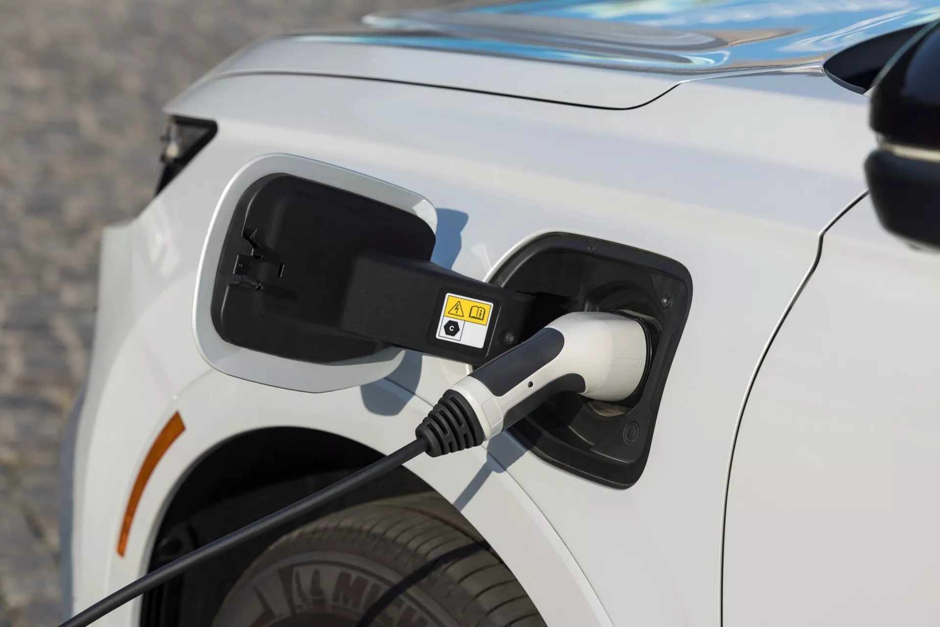 Zbliżenie na obszar portu ładowania białego elektrycznego pojazdu Honda CR-V 2023, pokazujące podłączony kabel i szczegóły takie jak otwarta klapka, naklejki ostrzegawcze i widok na tylną oponę.