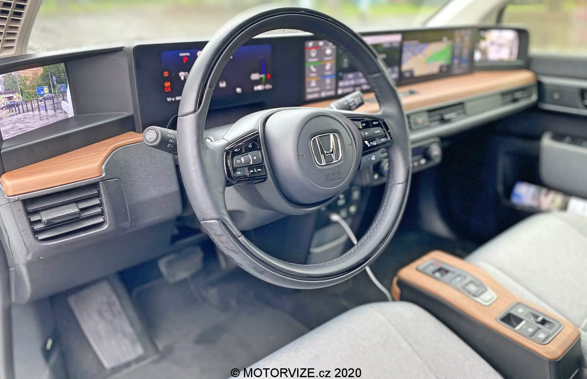 Wnętrze Hondy E 2020 pokazujące stronę kierowcy z naciskiem na kierownicę i nowoczesną deskę rozdzielczą. Zbliżenie podkreśla logo Honda na kierownicy, która posiada zamontowane sterowanie do multimediów i tempomatu. Wydłużone, ciągłe ekrany służą jako cyfrowy zestaw wskaźników pokazujący prędkość oraz system infotainment z mapą nawigacji. Widoczna drewniana wykończenia dodają luksusu, a jasne tapicerowane fotele sugerują komfort i styl. Zaawansowane funkcje jak panele dotykowe, przycisk uruchamiania i zapłon bezkluczykowy uwydatniają technologicznie zaawansowaną kabinę.
