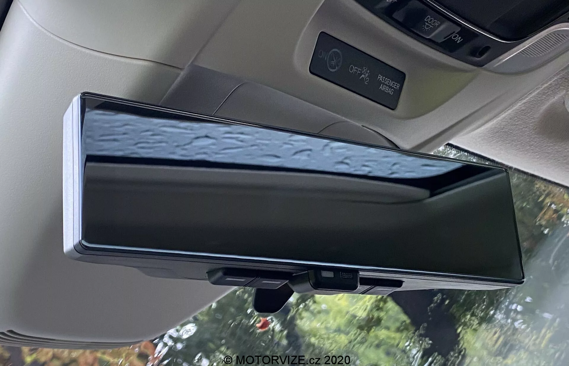 Widok wnętrza Hondy E 2020 z cyfrowym lusterkiem wstecznym. Elektroniczne urządzenie, umieszczone w tradycyjnym obudowaniu lusterka wstecznego, jest głównym tematem, pokazującym transmisję na żywo z tylnej kamery. Lustro pokazuje odbicia drzew z kroplami wody i oferuje lepszą widoczność w porównaniu z tradycyjnymi lusterkami. Zdjęcie ukazuje podsufitkę samochodu, światła kabinowe, osłonę przeciwsłoneczną i panel sterowania szyberdachem, podkreślając technologię zaawansowanych systemów wspomagania kierowcy (ADAS).