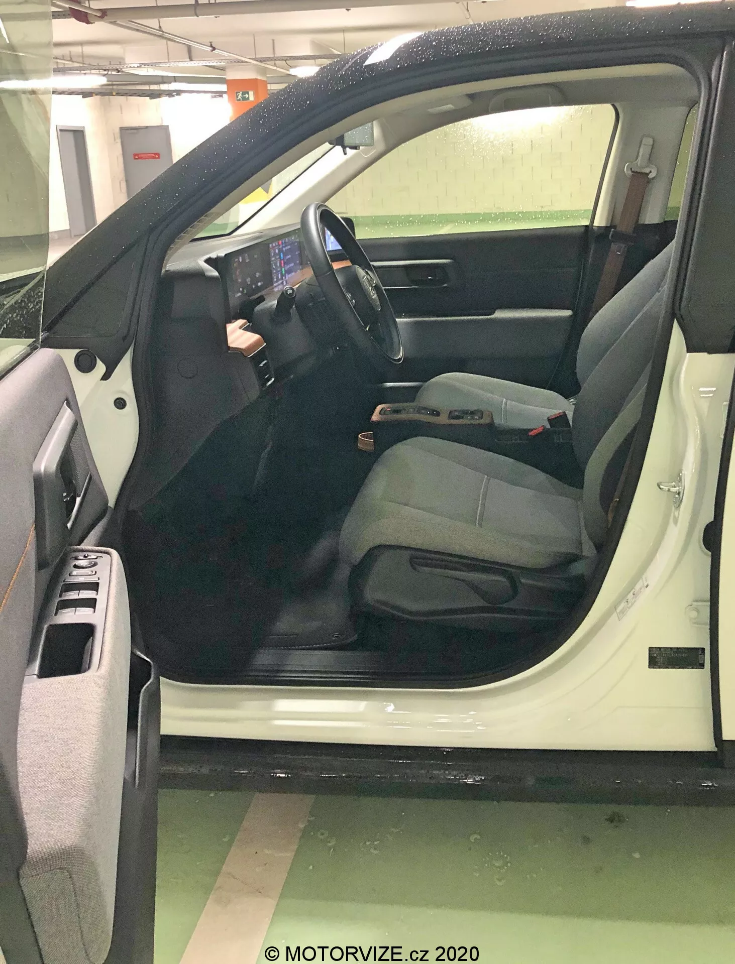 Zbliżony widok otwartych drzwi kierowcy samochodu z nieco podwyższonego kąta, skupiający się na nowoczesnym wnętrzu zorientowanym na technologię. Duży, wyraźny centralny ekran dotykowy, minimalna liczba fizycznych przycisków i zamontowane na kierownicy przyciski podkreślają zaawansowane systemy informacyjno-rozrywkowe i kontrolne. Fotel kierowcy sugeruje komfort z regulowanym zagłówkiem. Szczegóły obejmują schowki w drzwiach, kratkę głośnika, sterowanie szybami elektrycznymi, pasy bezpieczeństwa, dźwignie regulacyjne i dywaniki, sygnalizując nowszy, zadbany model.