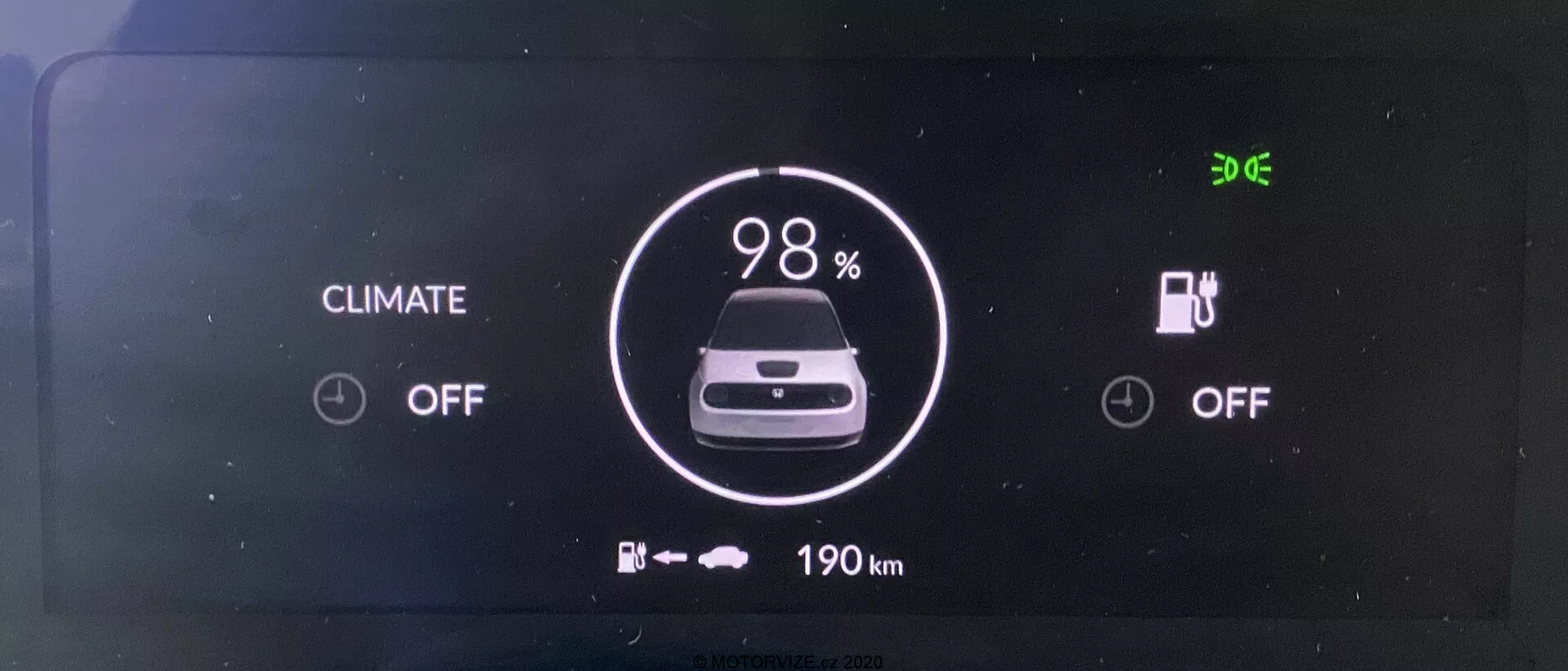 Cyfrowy panel instrumentów elektrycznego pojazdu Honda e 2020. Ekran, widziany od przodu, wyraźnie pokazuje wskaźnik baterii na poziomie 98% naładowania, ikonę auta i '190 km' pozostałego zasięgu poniżej. Statusy systemu kontroli klimatu i ładowania są obydwa 'OFF'. Czas '9:06' jest wyświetlany w lewym górnym rogu.