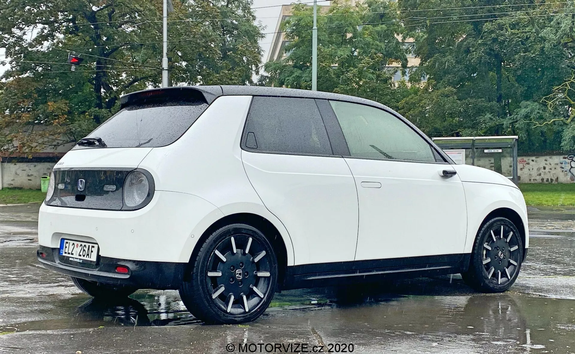 Obraz przedstawia boczny i tylny widok białego nowoczesnego hatchbacka Honda e 2020, sfotografowanego z kąta trzy-czwarte. Mokra powierzchnia i odbicia światła sugerują niedawną ulewę. Widoczne cechy obejmują profile boczne i tylne, dwoje drzwi, kanciasty kształt z wyraźnymi nadkolami, duże czarne koła z wieloramiennym sportowym wzorem, małe tylne okno z wycieraczką, tylny spojler dachowy, wyraźne tylne światła obejmujące boki, centralnie umieszczoną tylną tablicę rejestracyjną oraz charakterystyczny znak na tylnej klapie. Cechy technologiczne to m.in. elementy aerodynamiczne, sportowy design kół, nowoczesne tylne światła prawdopodobnie z technologią LED oraz poprawiona widoczność tylnej szyby.