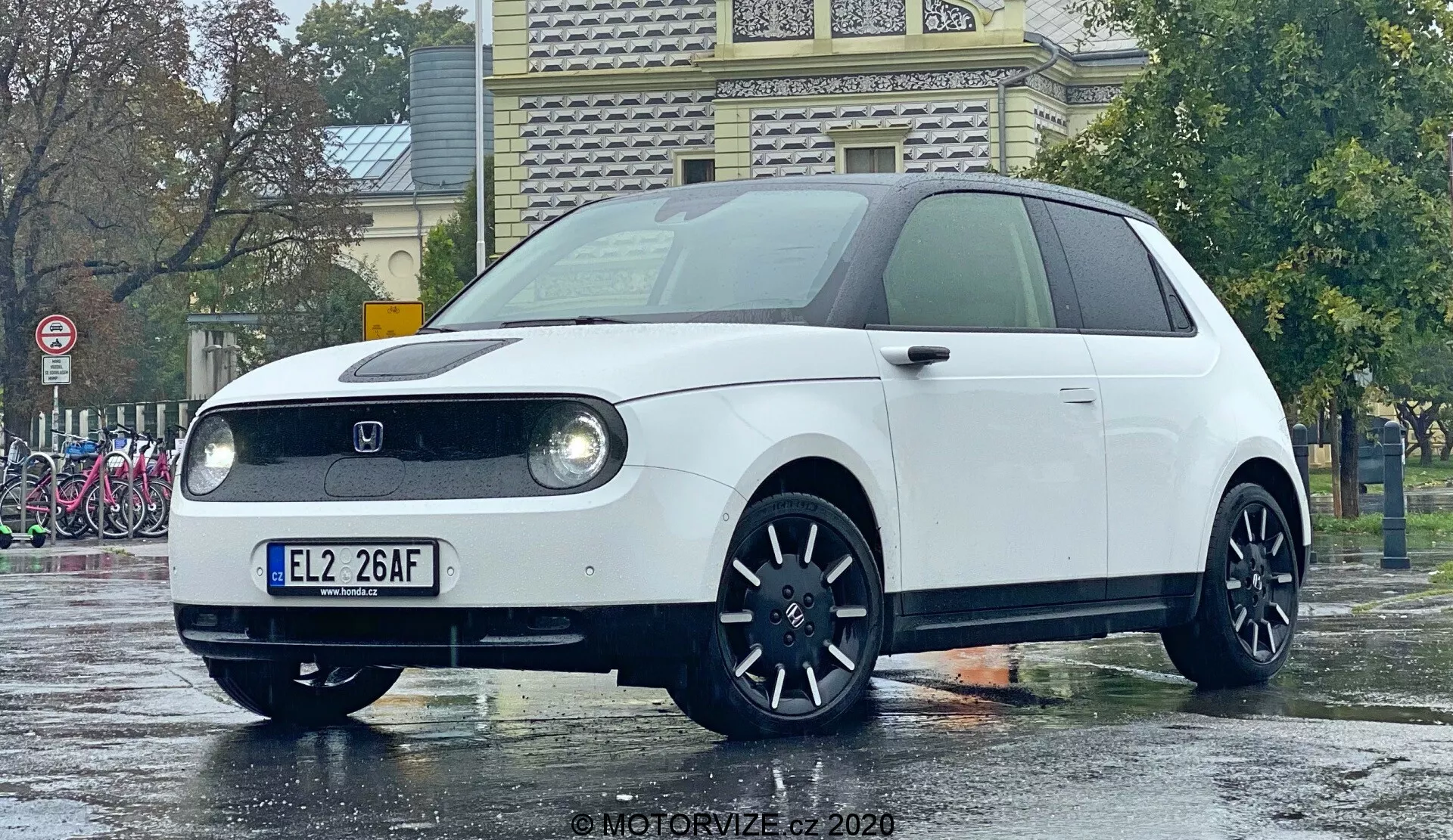 Honda e 2020 sfotografowany pod niskim kątem, podkreślający przedni koniec i częściowy profil boczny, wyróżniający okrągłe reflektory LED w prostokątnych obudowach, zamkniętą kratkę z przodu typową dla pojazdów elektrycznych, centralnie umieszczoną odznakę producenta, ciemne, wieloramienne felgi i minimalistyczny, retro-nowoczesny design. Kompaktowy hatchback stoi na mokrym podłożu odbijającym deszczowe otoczenie, sprawiając, że jego czyste linie i kolor wydają się bardziej żywe.